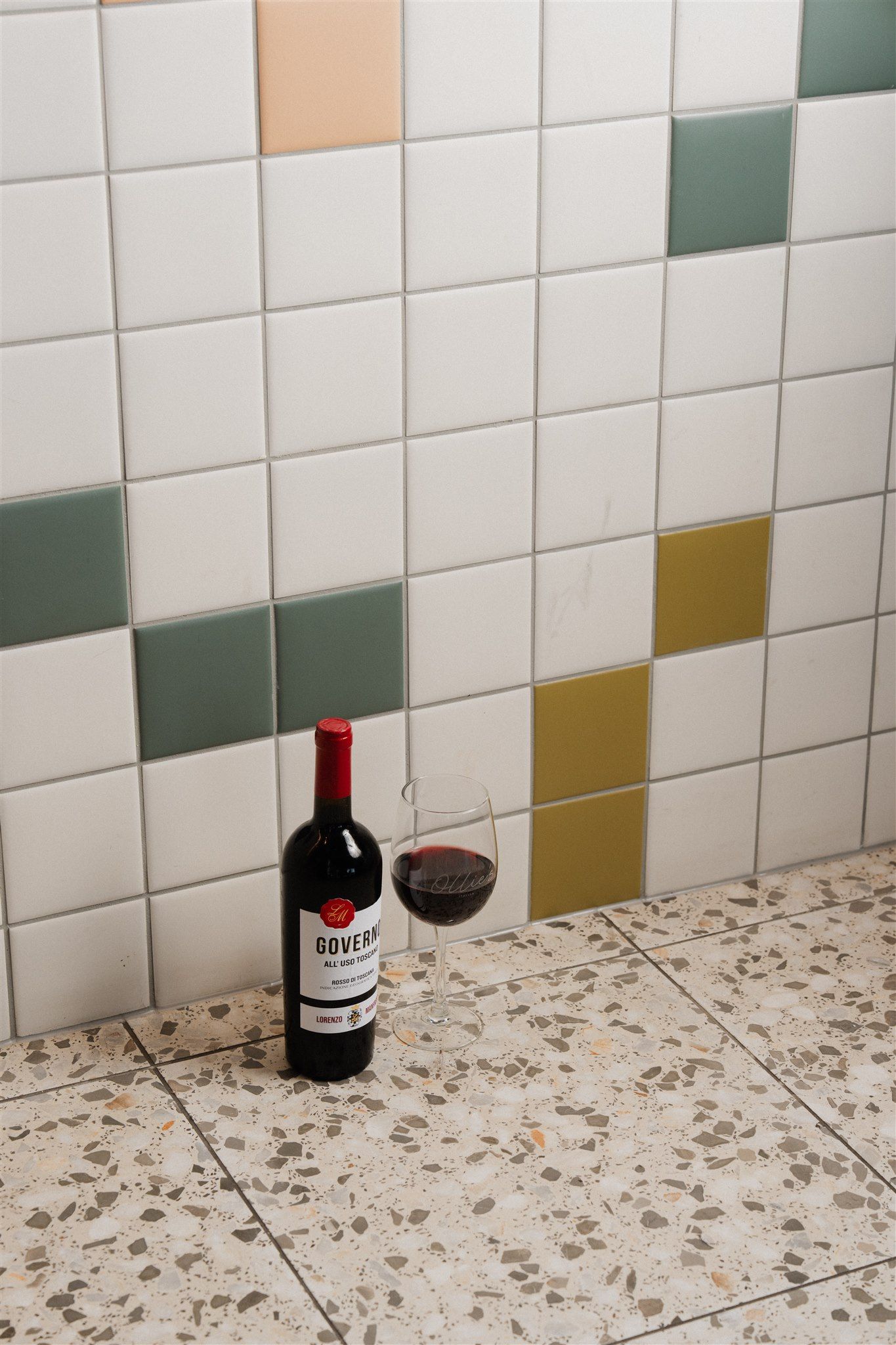 Wine bottle and filled glass placed on terrazzo flooring against a tiled wall with muted green, ochre and blush accent tiles at Ollie’s Italian.