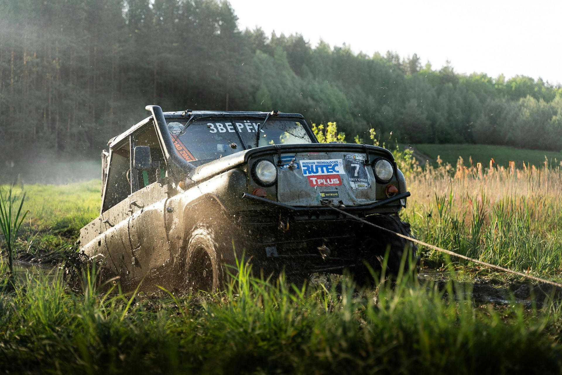 An off-road vehicle stuck in muddy marsh being pulled by a rope; forest in background.