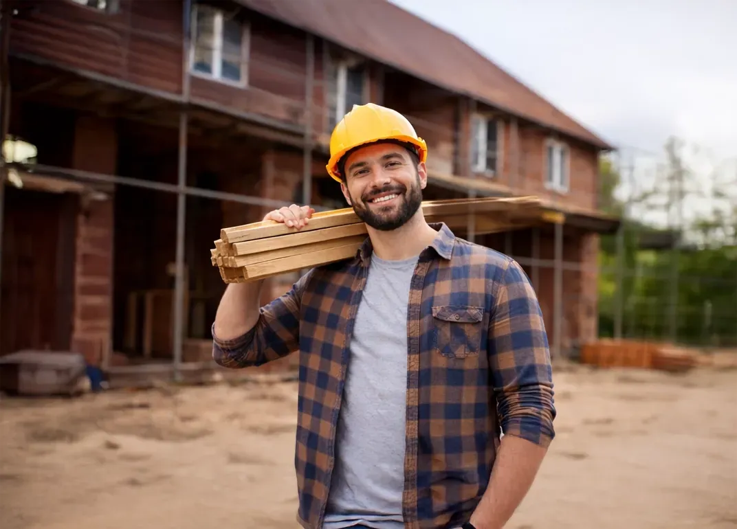 Construction worker holding wood on shoulder, smiling, wearing hard hat in front of a house under construction.