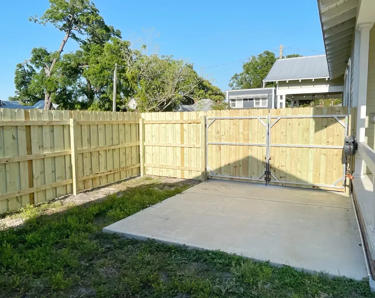 A fenced-in backyard with a concrete patio and gate, light brown fence, green grass, and a clear blue sky.