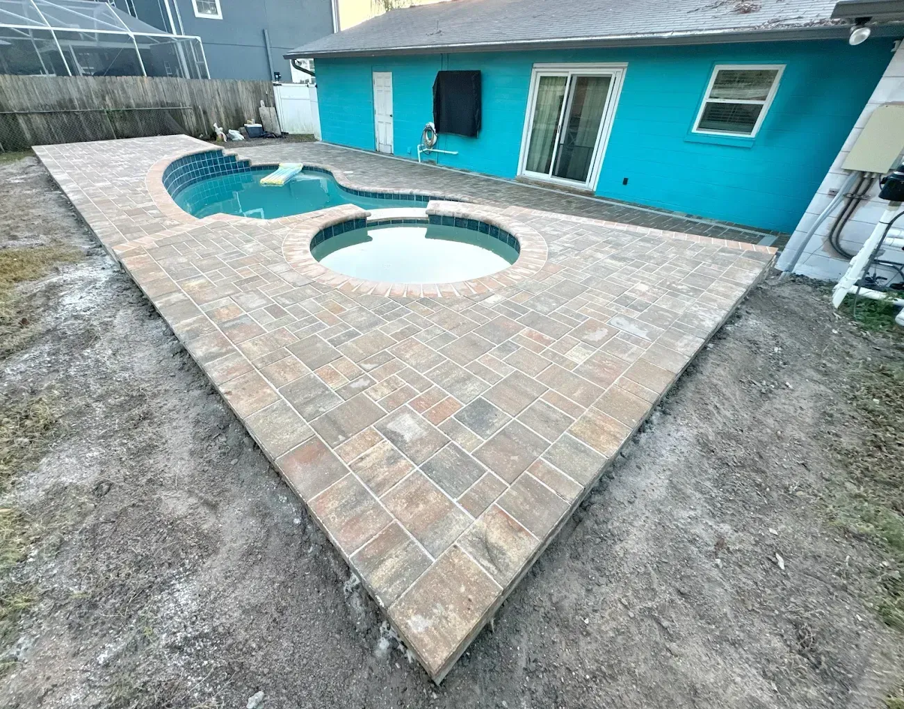 Brick patio surrounds a pool and jacuzzi next to a turquoise house.