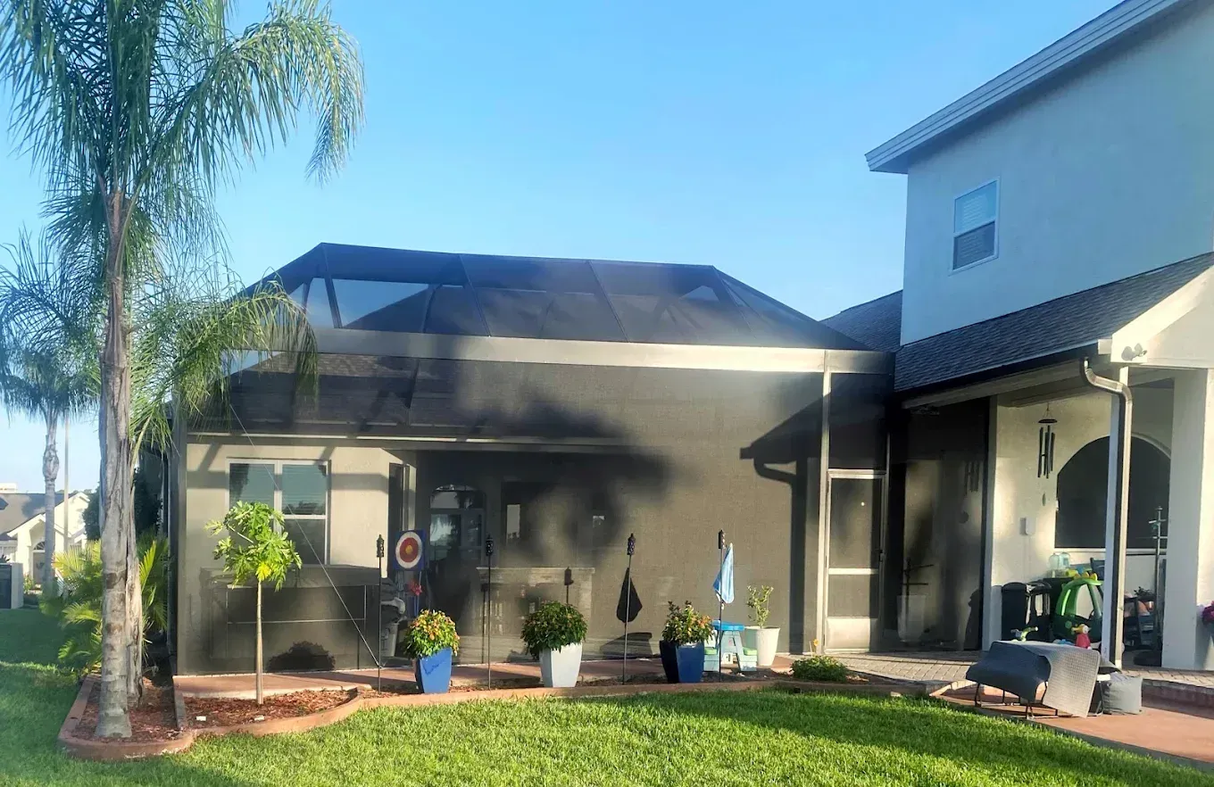 Backyard patio with screened enclosure, plants, and a two-story house under a blue sky.