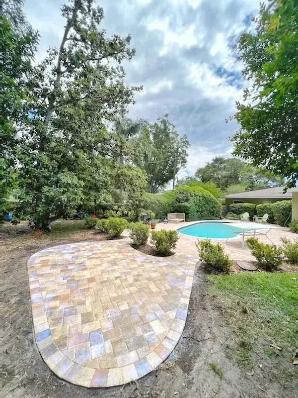 Brick patio curves towards a turquoise pool in a backyard with green bushes and trees, under a cloudy sky.