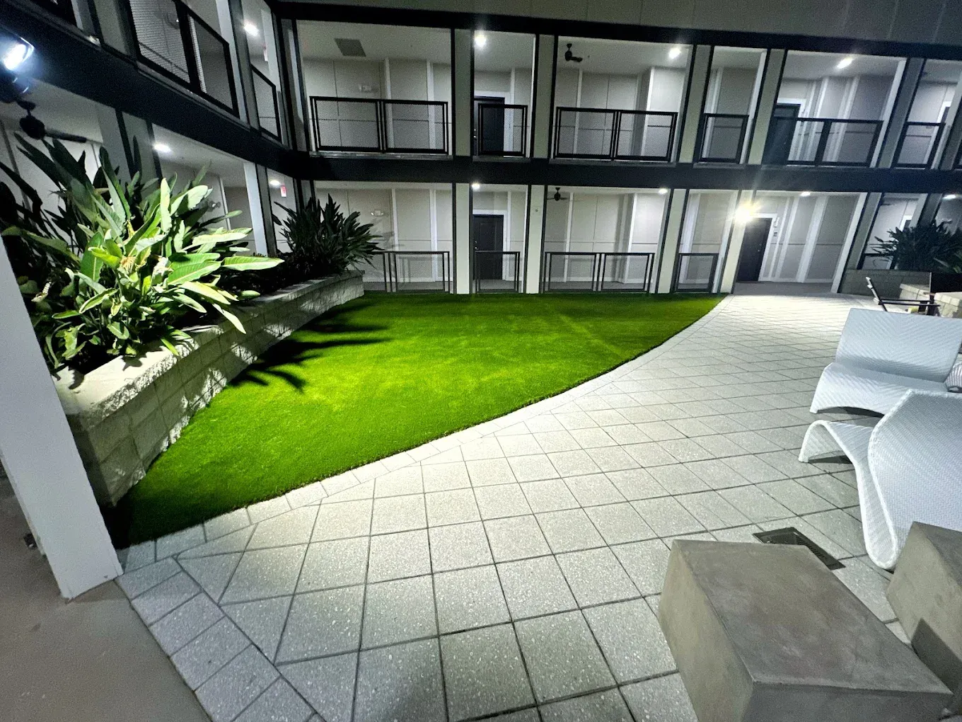 Courtyard with artificial grass, paved walkways, white benches, and two-story building with balconies at night.
