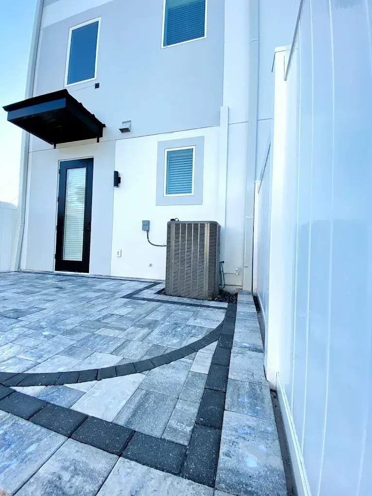 A modern home exterior with a paved patio, AC unit, and a black door under an awning.