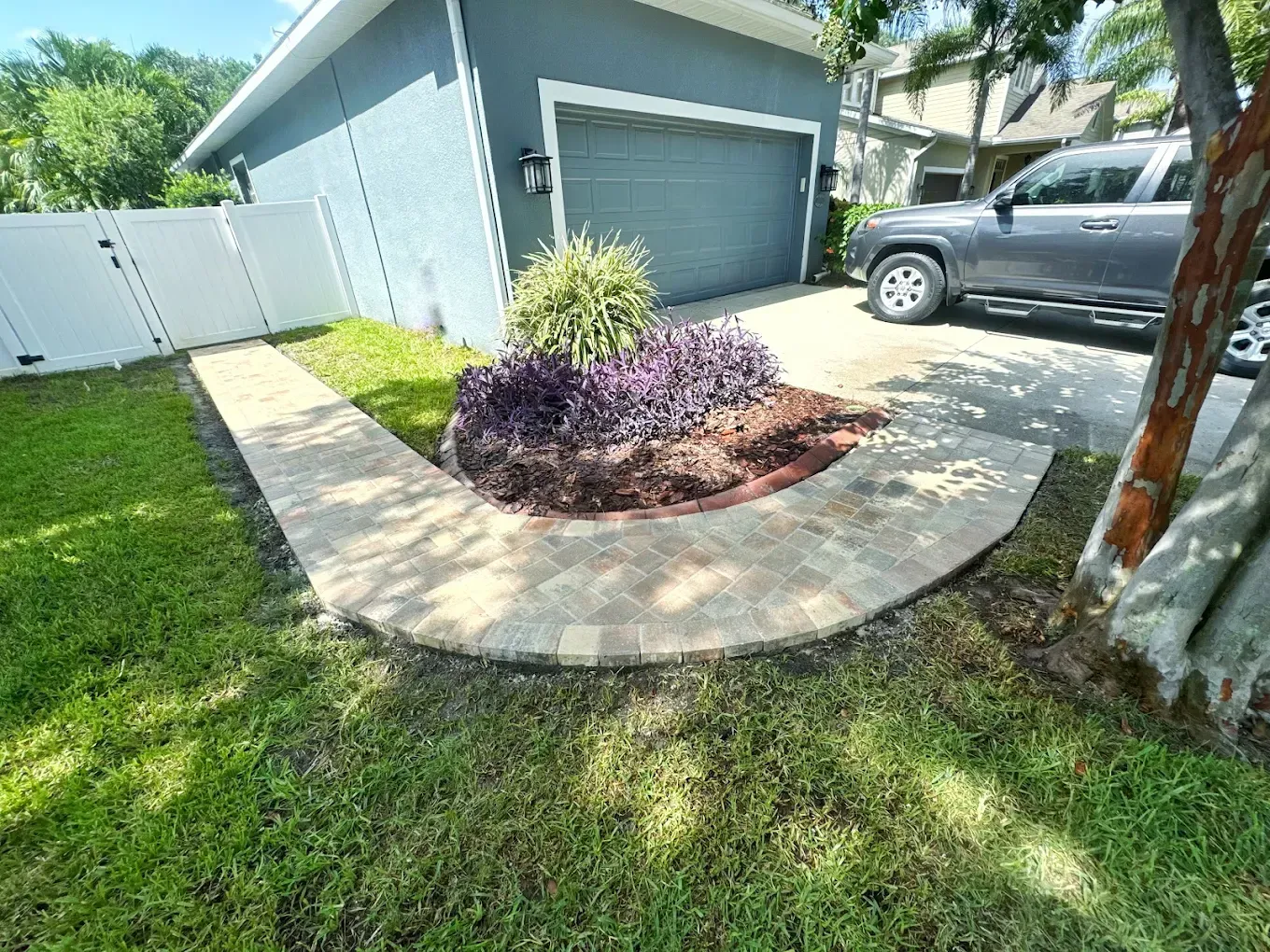 Brick walkway curves around a landscaped flower bed with purple foliage, next to a garage and a truck.