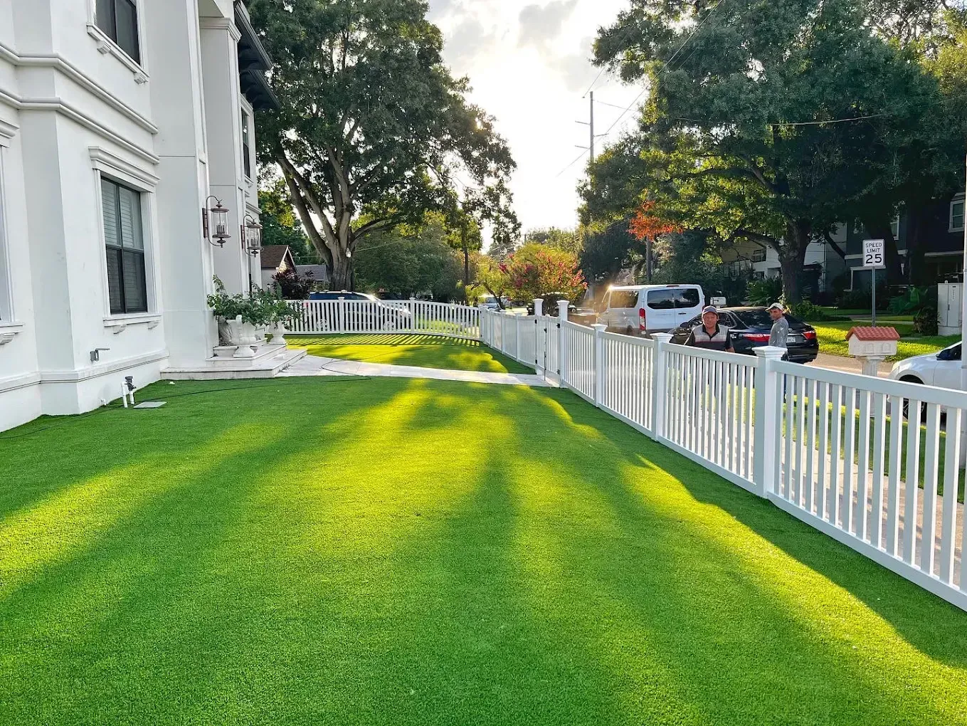 Lush green lawn with white picket fence, next to a white building. Cars and trees line the street.
