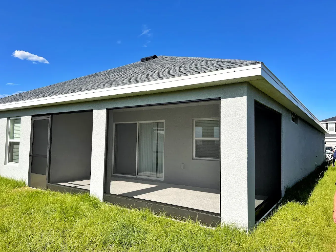 Back patio with screened enclosure. Grey walls, concrete floor, and green grass under a blue sky.