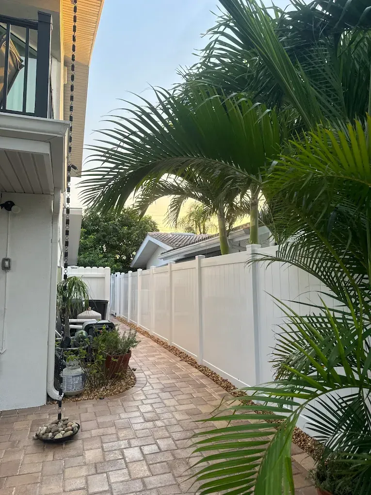 Pathway between a white building and fence, with palm trees and brick paving.