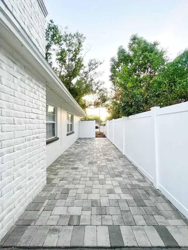 Brick walkway between a white brick building and a white fence, with trees in the background.