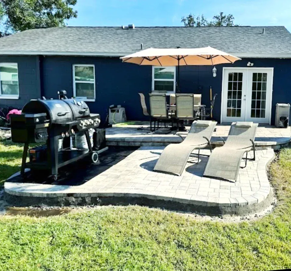 Backyard patio with grill, lounge chairs, dining set, and umbrella in front of a blue house.