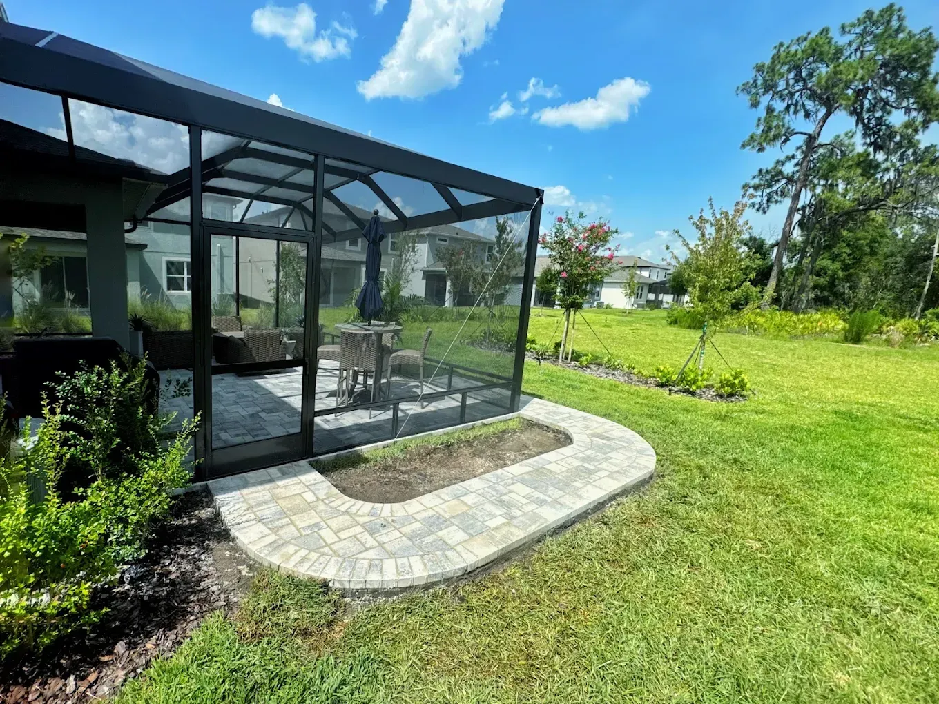 Screened patio with table and chairs, framed by a brick border and lawn, under a bright blue sky.