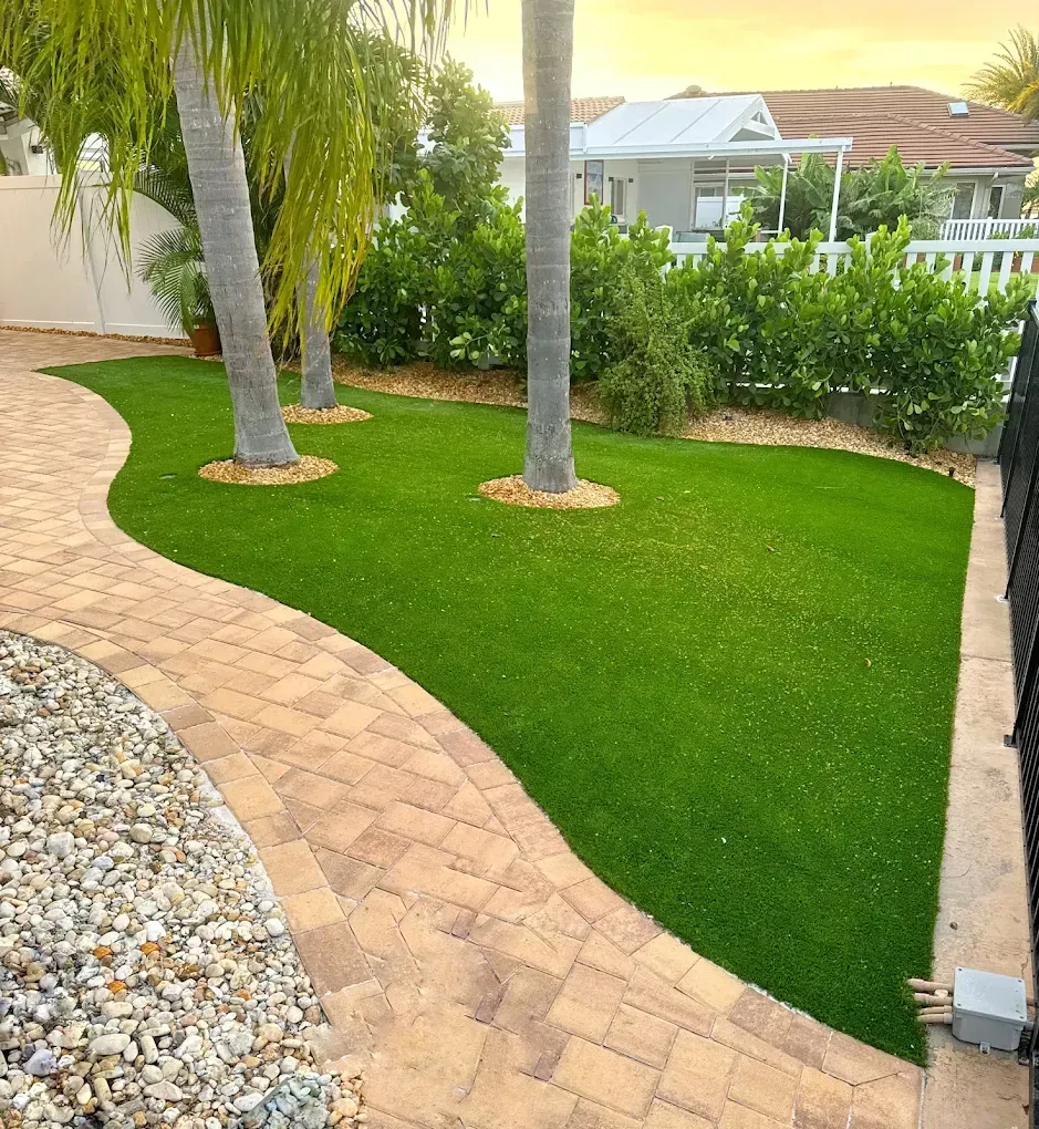 Brick pathway curves through vibrant green lawn with palm trees, bushes, and a house in the background.