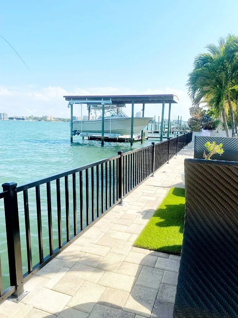 Waterfront view with black railing, dock with boat, green turf, blue water and sky.