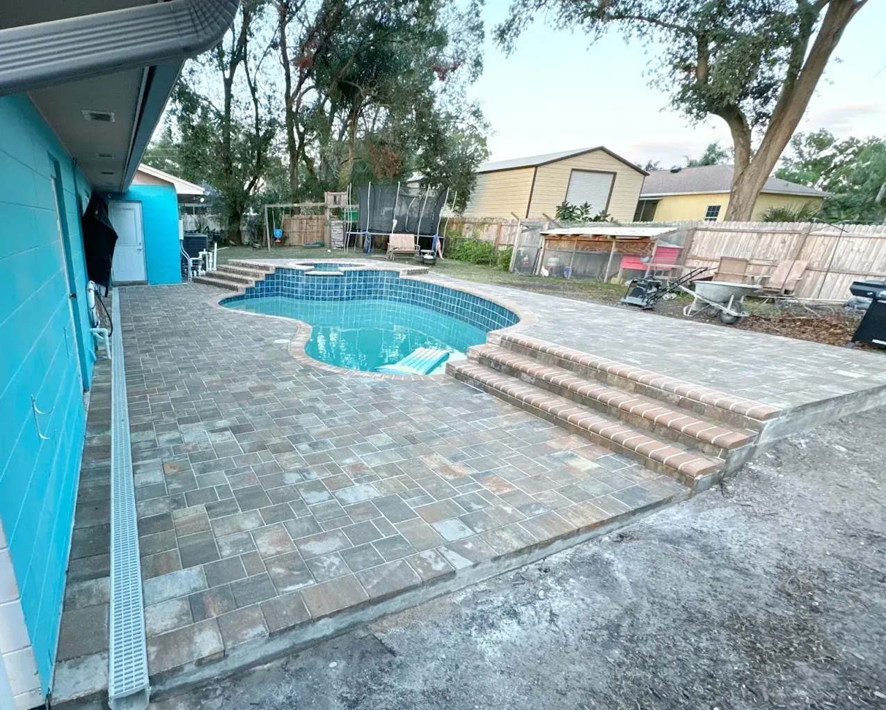 Backyard with pool, blue house, brick patio, steps leading to pool. Trees, fence, and shed in background.