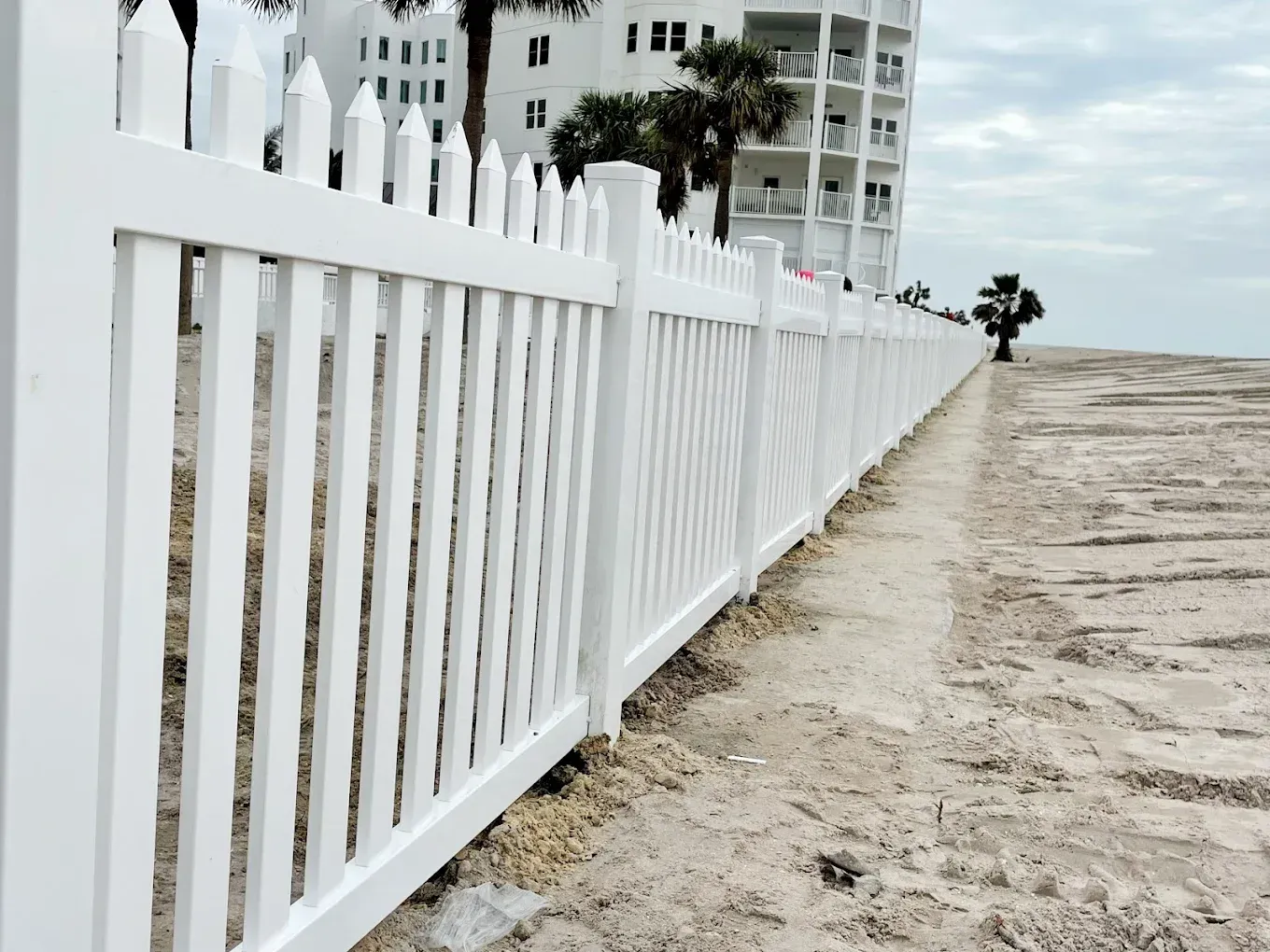 White picket fence along a sandy beach, with a building in the background.