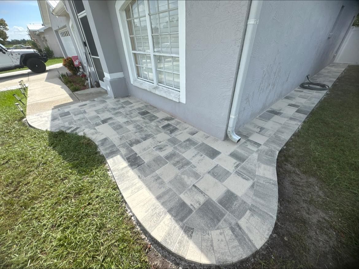 Brick paver walkway and patio, bordered by grass, leading to a light gray house with a window.
