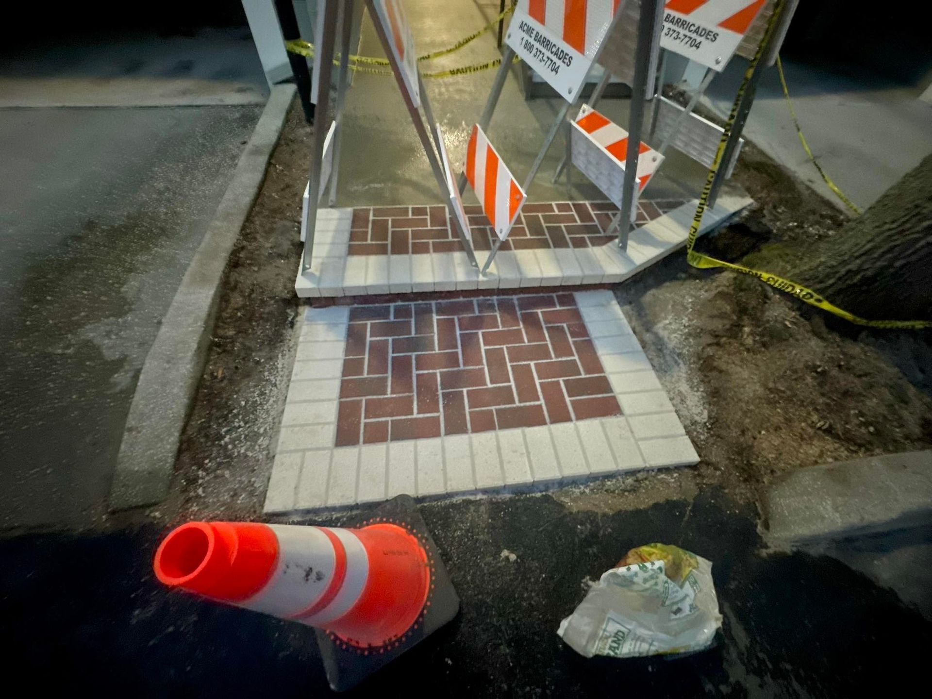 Sidewalk construction area, brick pattern with orange cone and caution signs, nighttime.