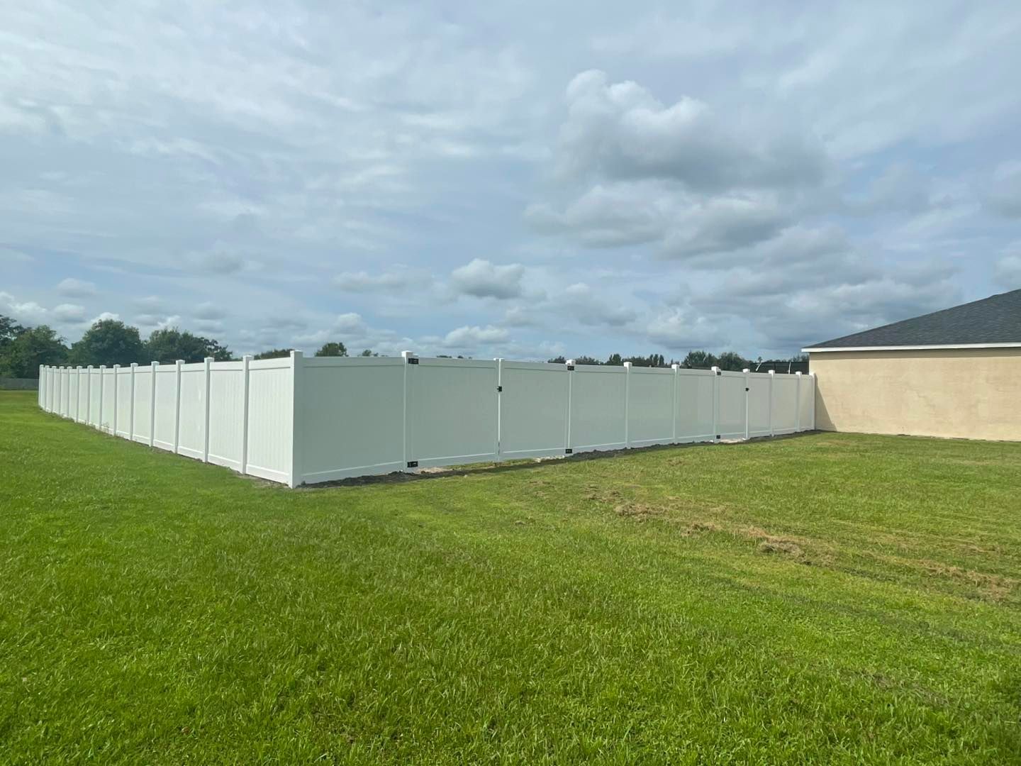 White vinyl fence surrounding a grassy yard, partially visible beside a beige building, cloudy sky.