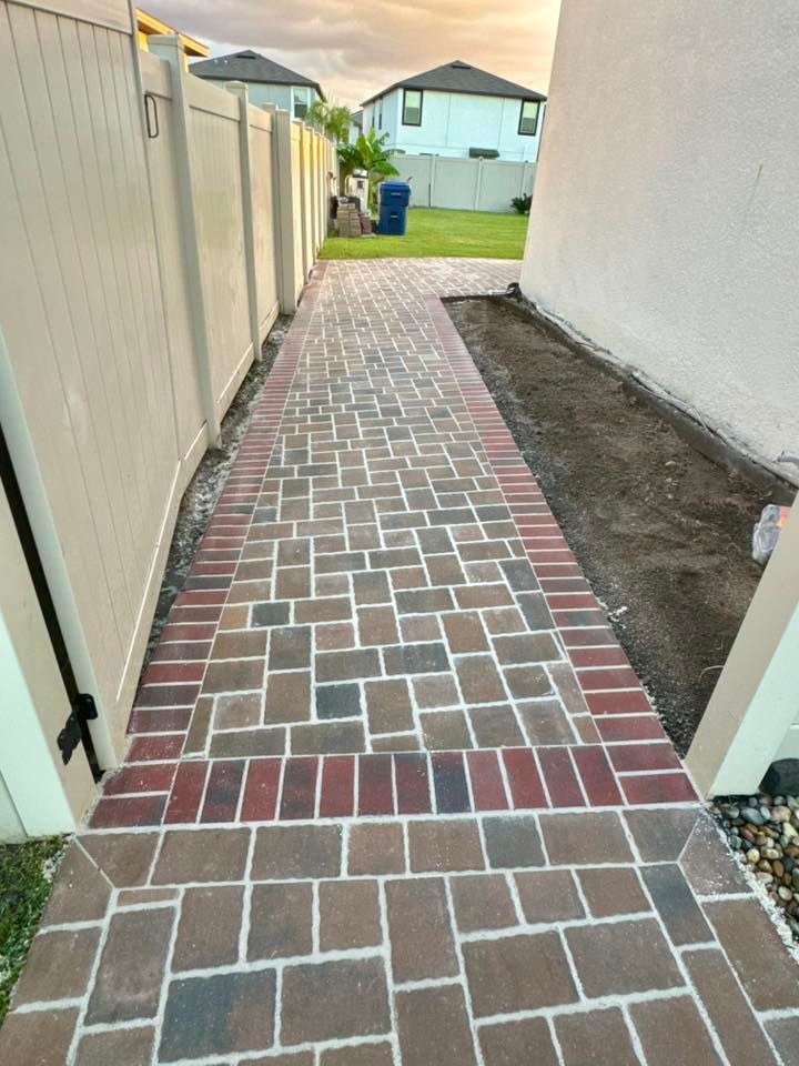 Brick walkway between a beige fence and a white wall; the pathway leads to a yard with homes.