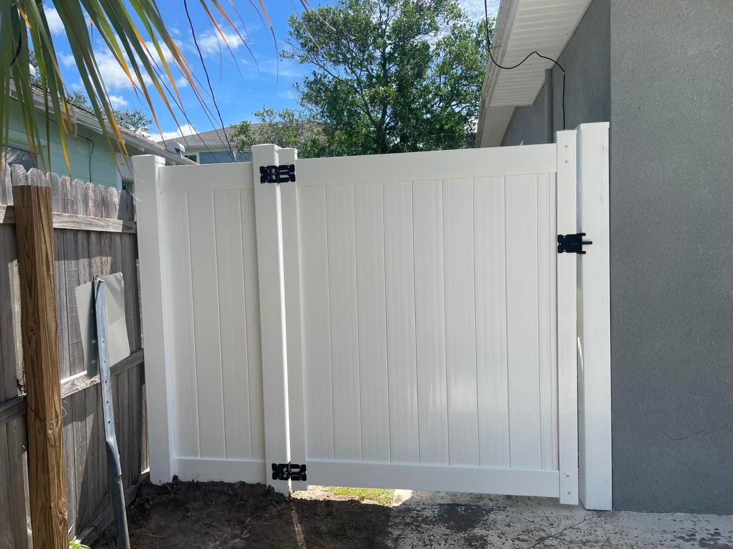 White vinyl fence gate, black hardware, against a gray wall and wooden fence.