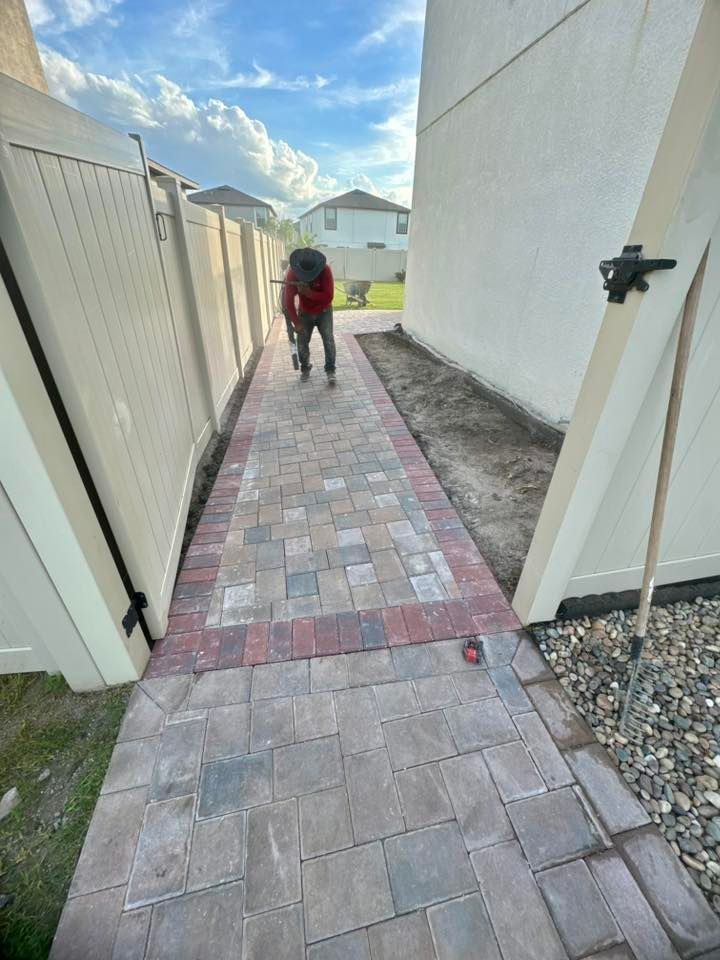 Person laying brick pavers in narrow pathway between fence and building.