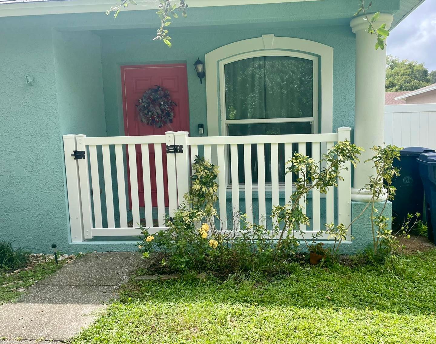 A turquoise house with a white picket fence. A red door is visible behind the fence. Green plants surround the fence.