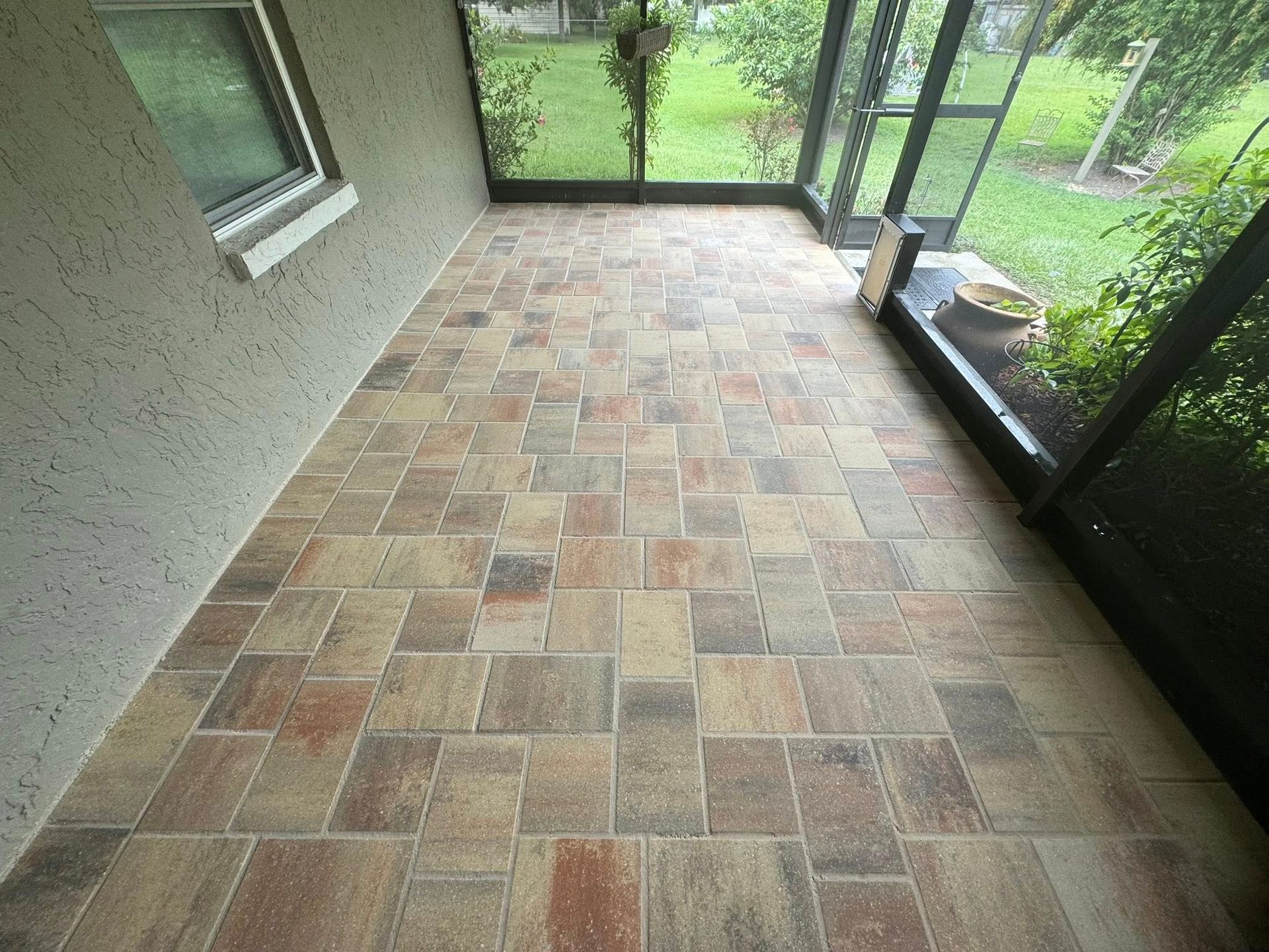 Screened-in patio with light and dark brown pavers; gray stucco wall; green foliage outside the screen.