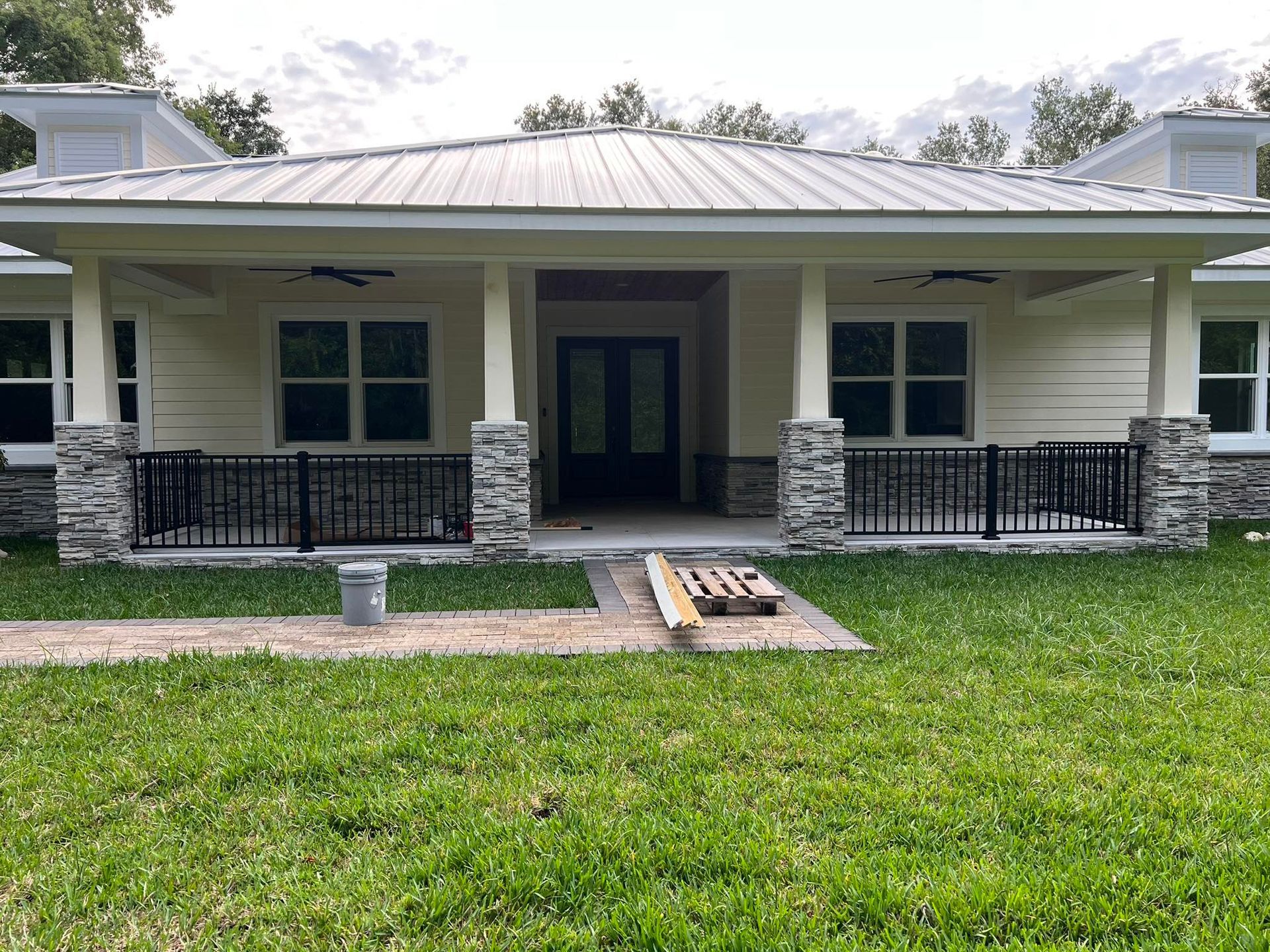 A light yellow house with a metal roof, stone pillars, black railings, and green grass.