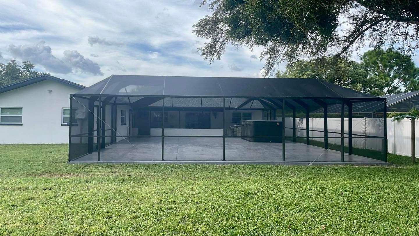 Screened-in patio with black mesh, concrete floor, and hot tub, attached to a white house, green lawn, cloudy sky.