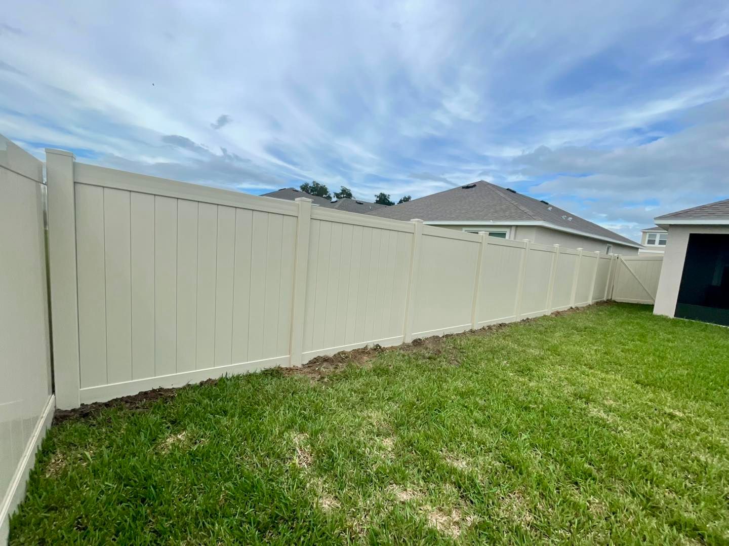Beige vinyl fence in backyard with green grass and cloudy sky.