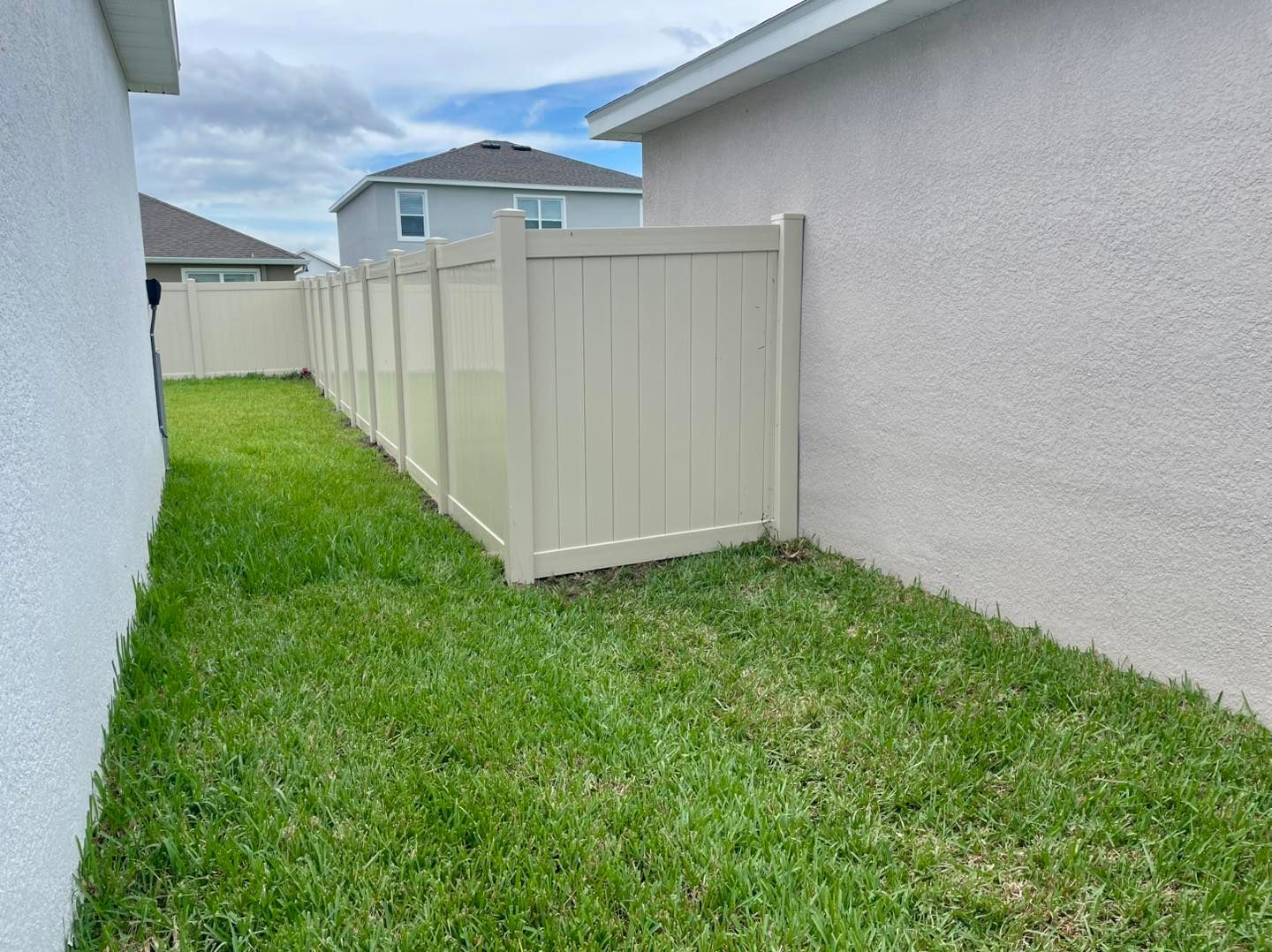 Lush green grass and a beige fence separate two light beige stucco houses on a cloudy day.
