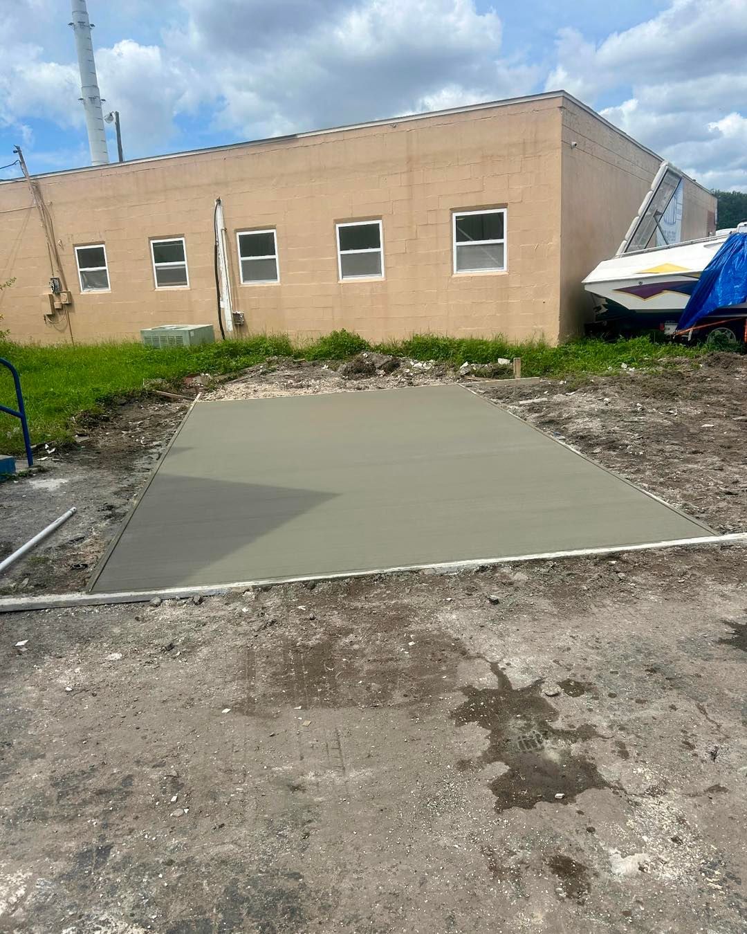 Newly poured concrete pad in front of a tan building, with a boat and cloudy sky in the background.