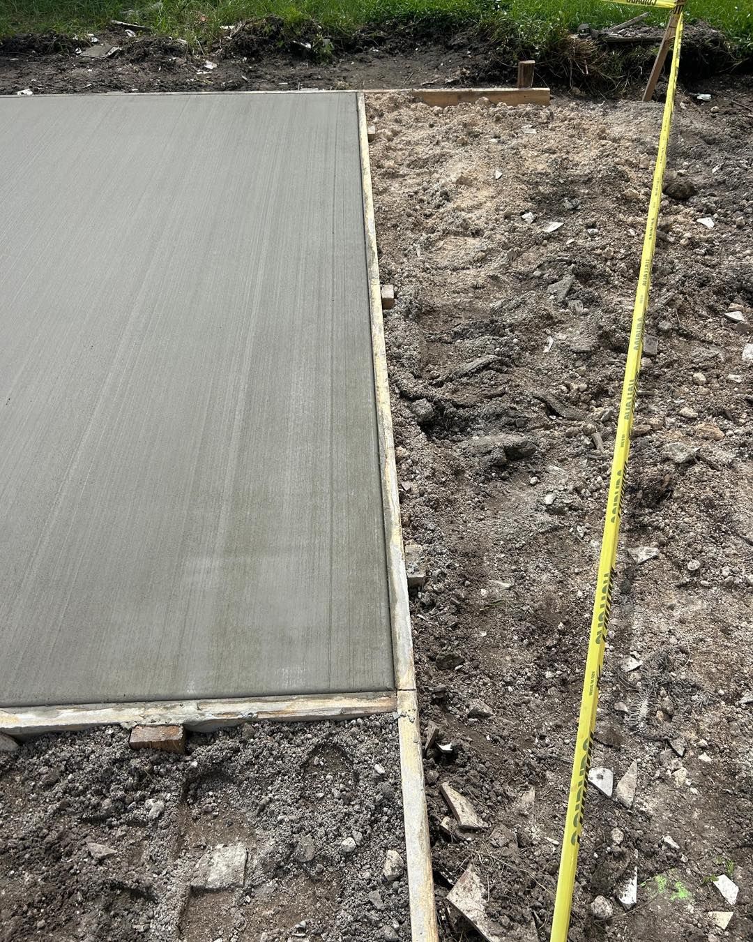 Freshly poured concrete slab next to unpaved ground, wooden formwork visible; yellow tape measure leans.