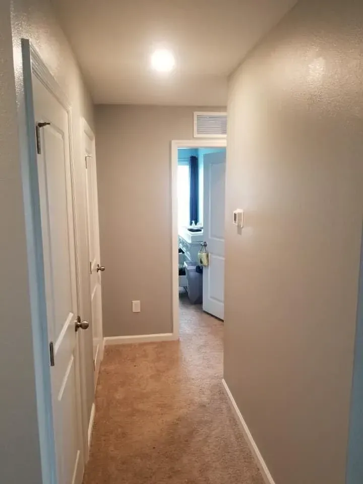Narrow hallway with beige walls, tan carpet, and white doors. A door at the end opens to a room with a blue window.