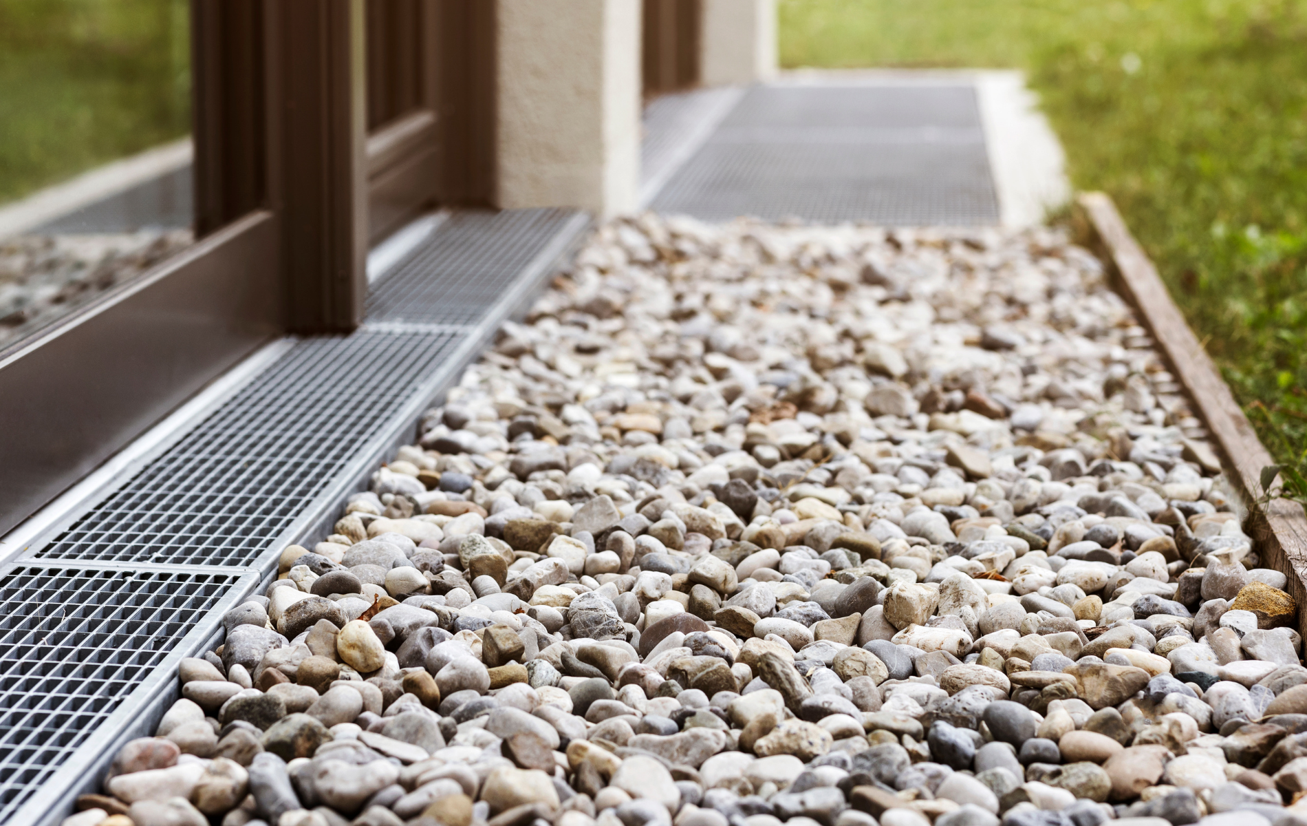 Gravel-filled bed next to a doorway with a drainage grate. Brown door frame, green grass on the right.