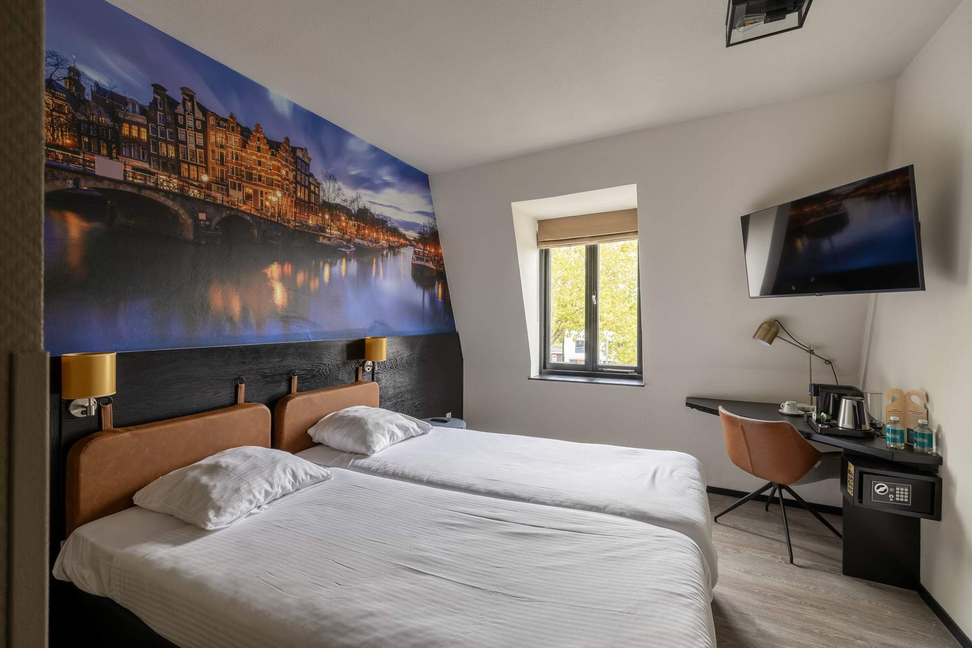 Hotelkamer met twee eenpersoonsbedden, bureau, tv en een muurschildering van een stadsgezicht.