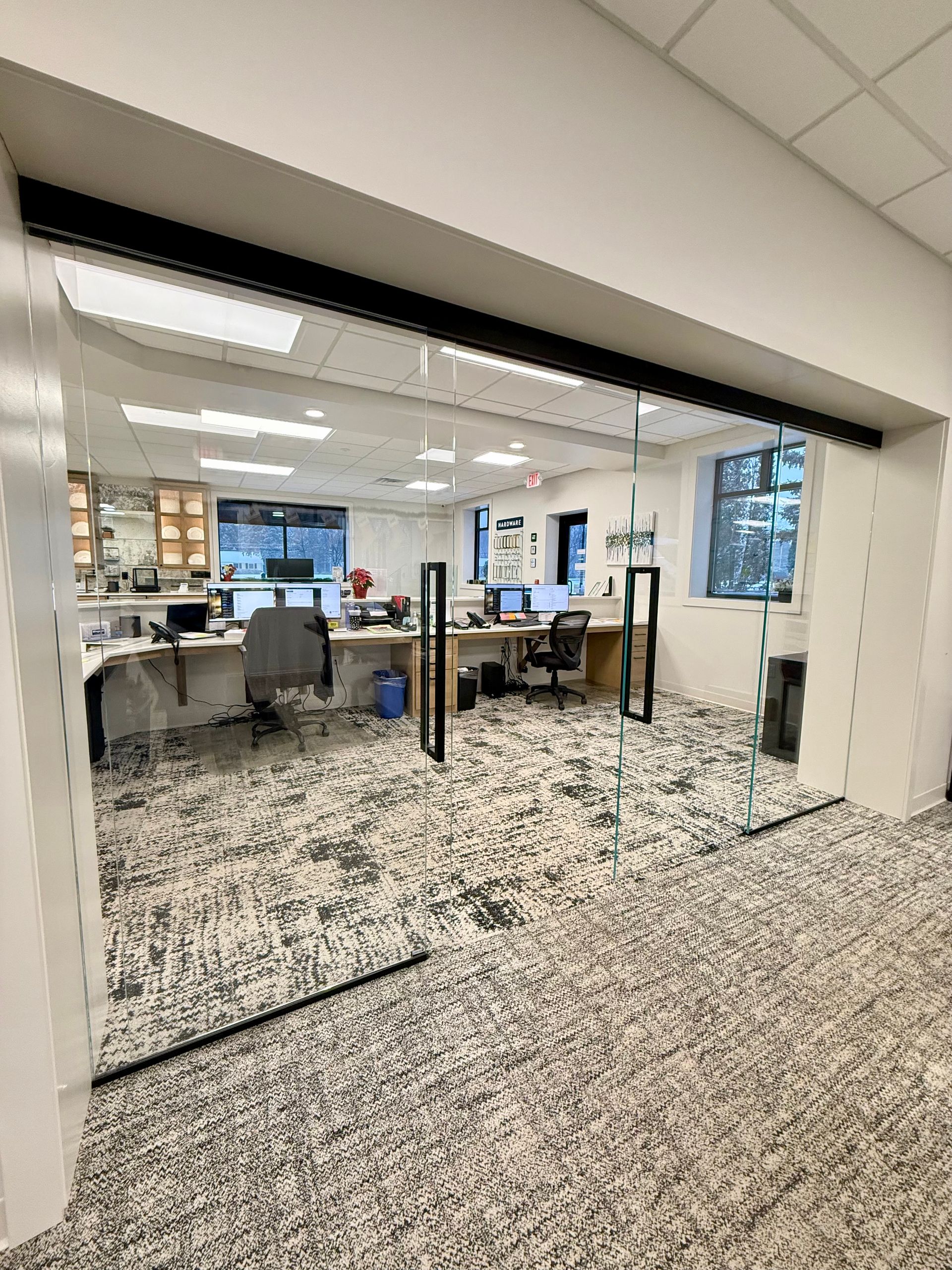 Open office space with glass doors. Desks and computers are visible inside. Light-colored carpet.