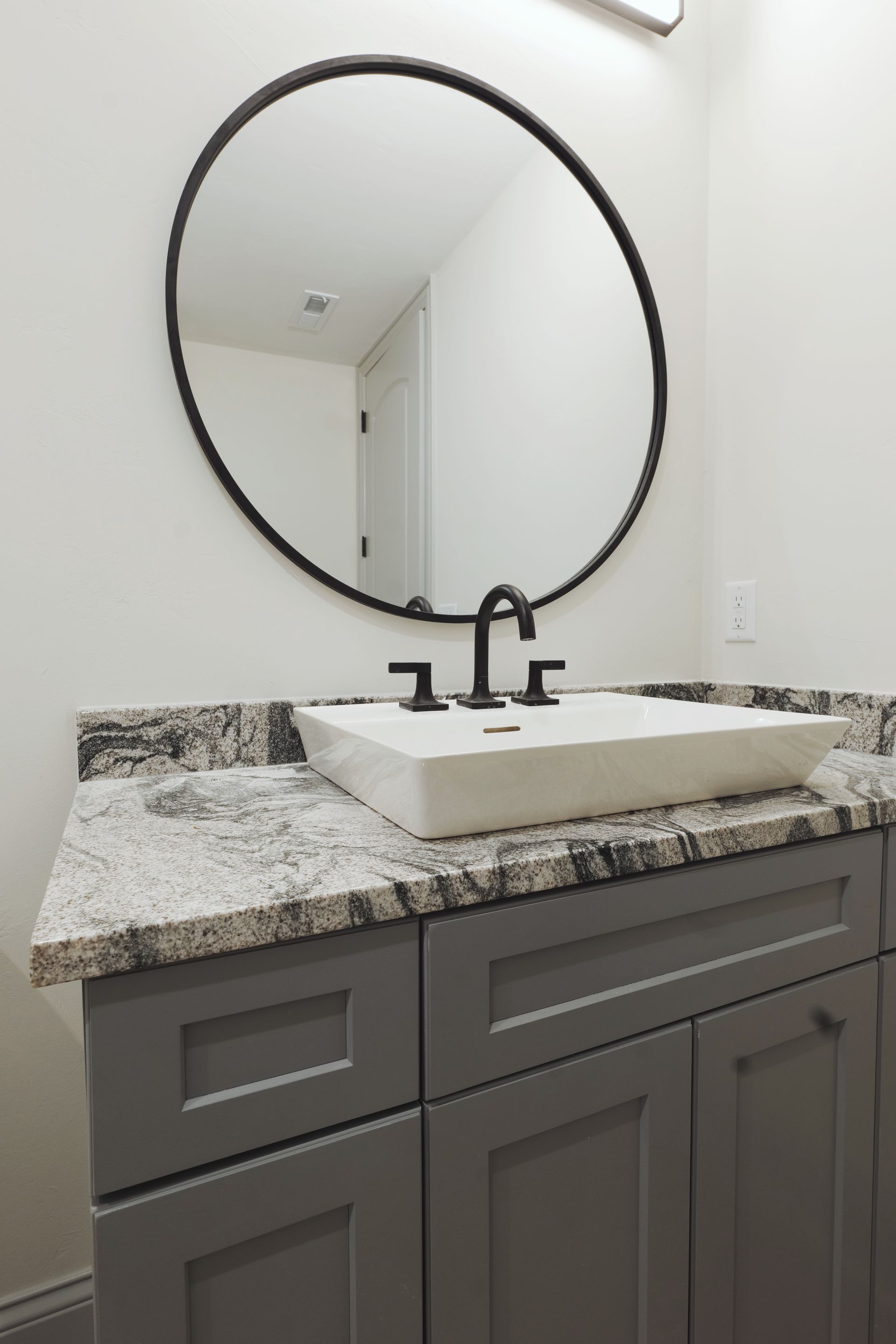 Bathroom vanity with a round mirror, gray cabinets, granite countertop, and white sink.