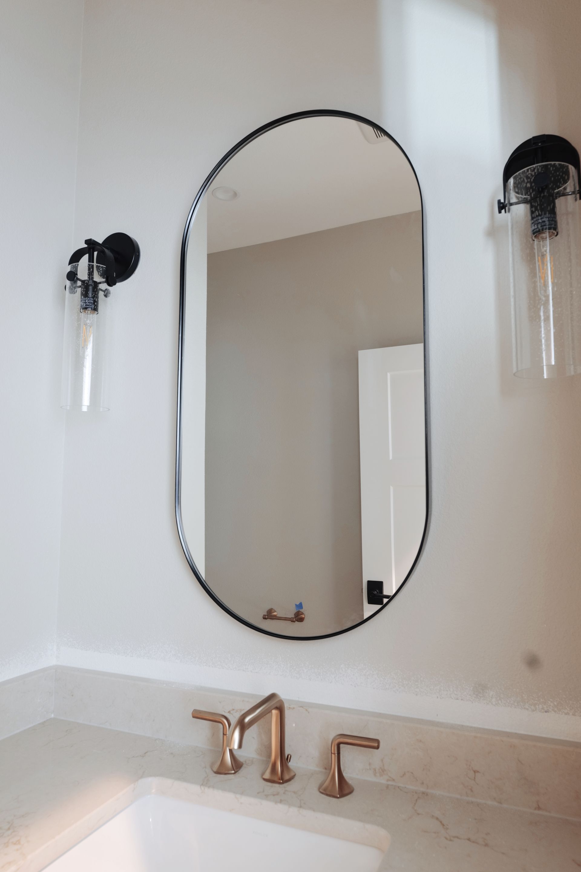 Bathroom with oval mirror, black frame, rose gold faucet, and glass light fixtures.