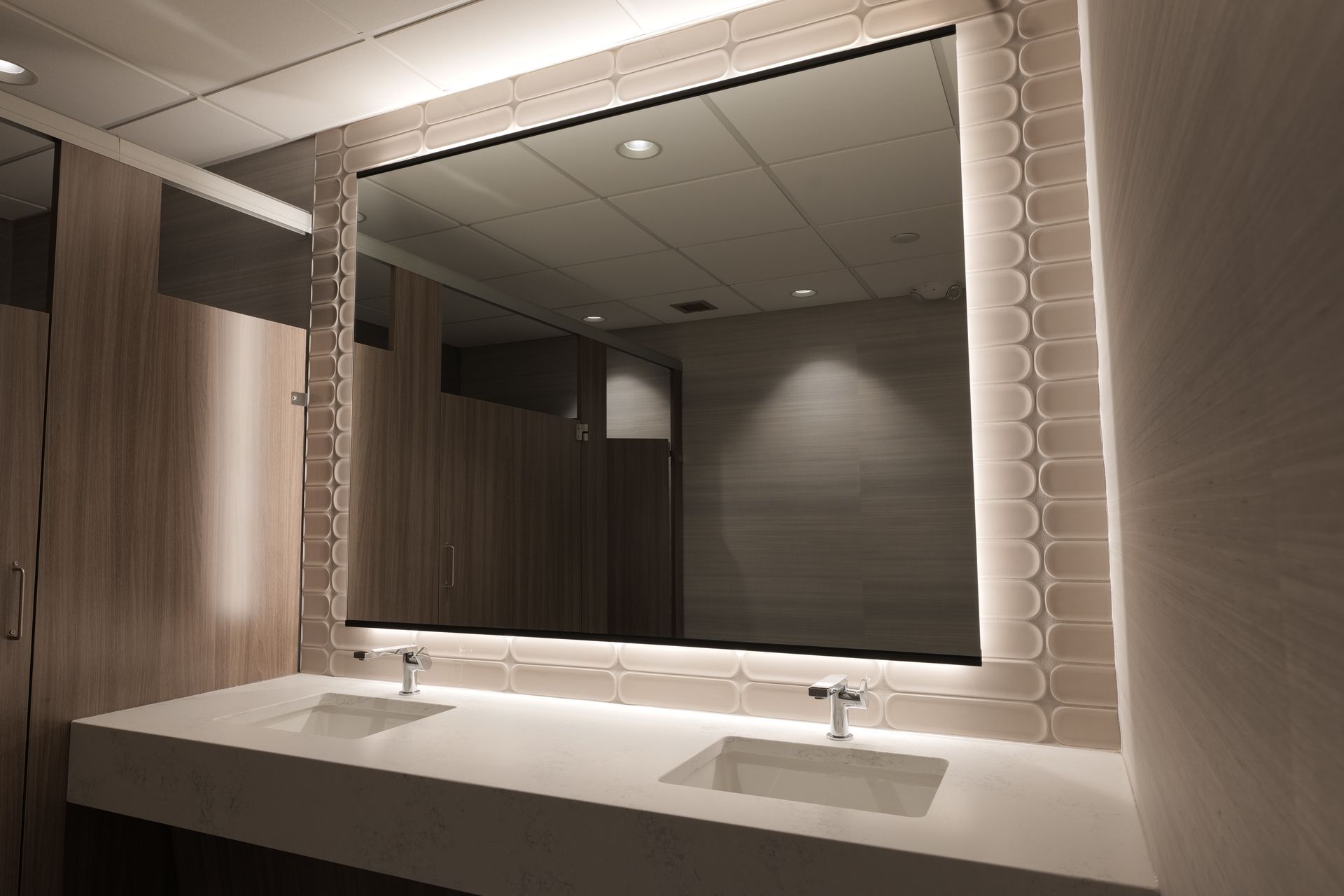 Bathroom with large illuminated mirror over a white countertop with two sinks and a row of stalls.