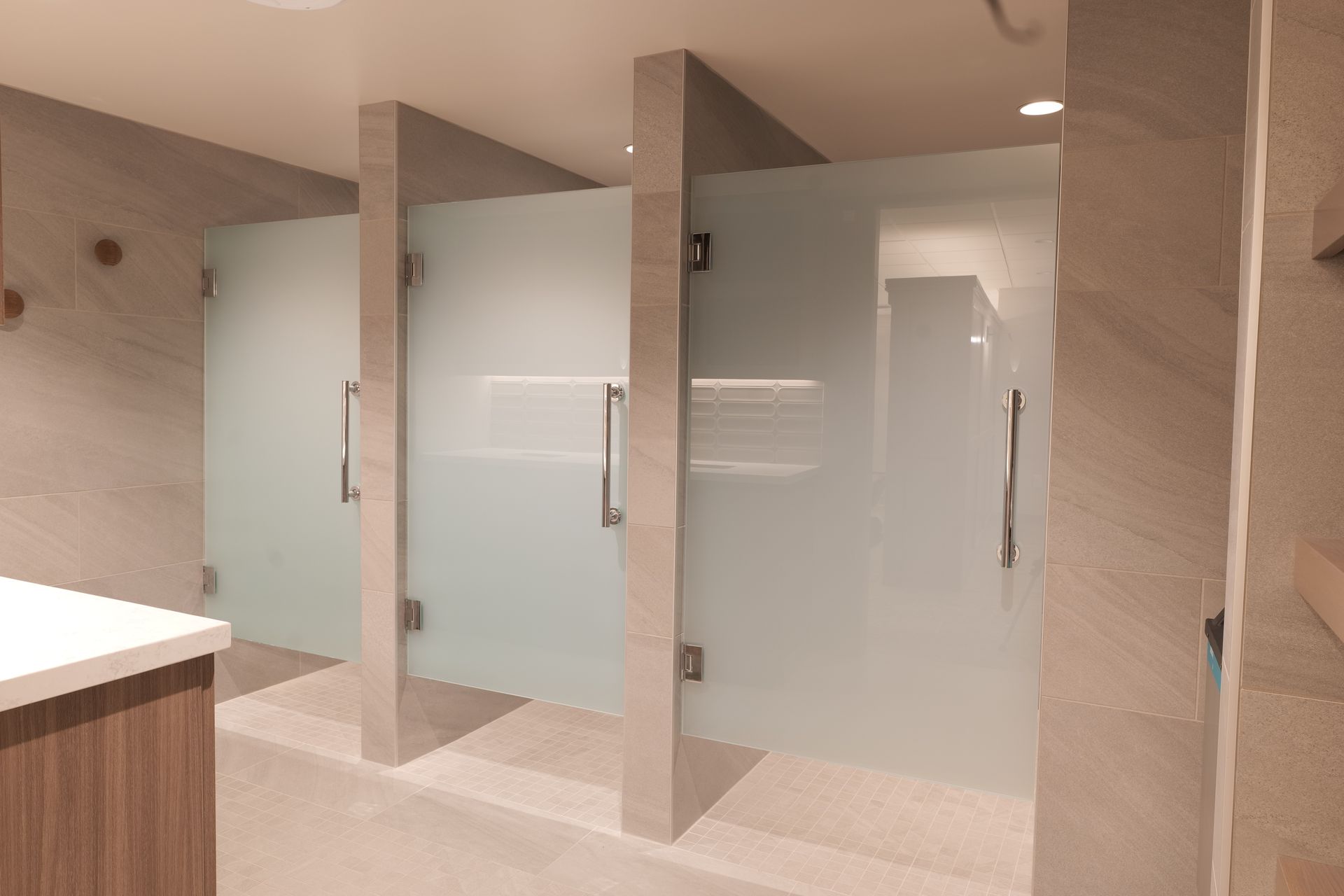 Three frosted glass shower stalls with silver handles in a light-colored tiled room.