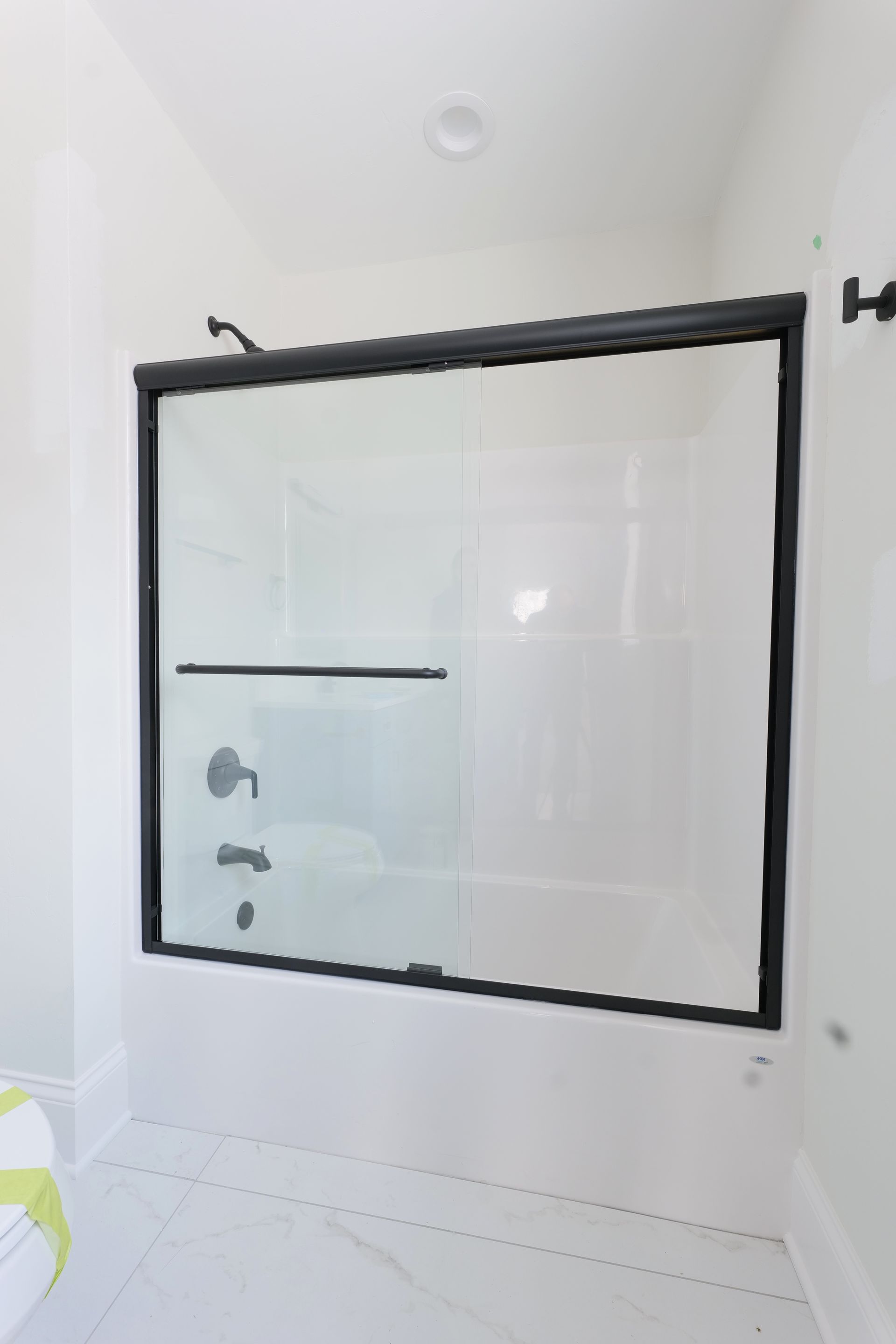 Black-framed shower door over a white tub in a bathroom with white walls and flooring.