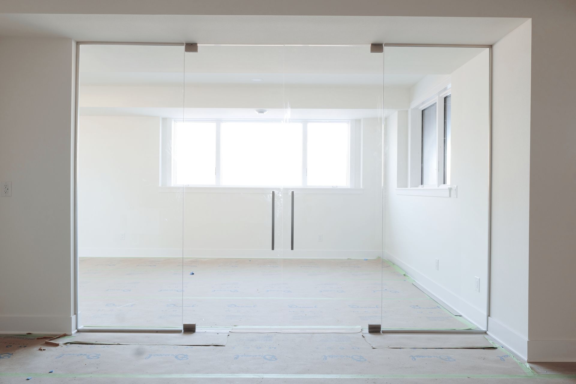 Glass doors opening to a white-walled room with a window, two handles, and unfinished flooring.