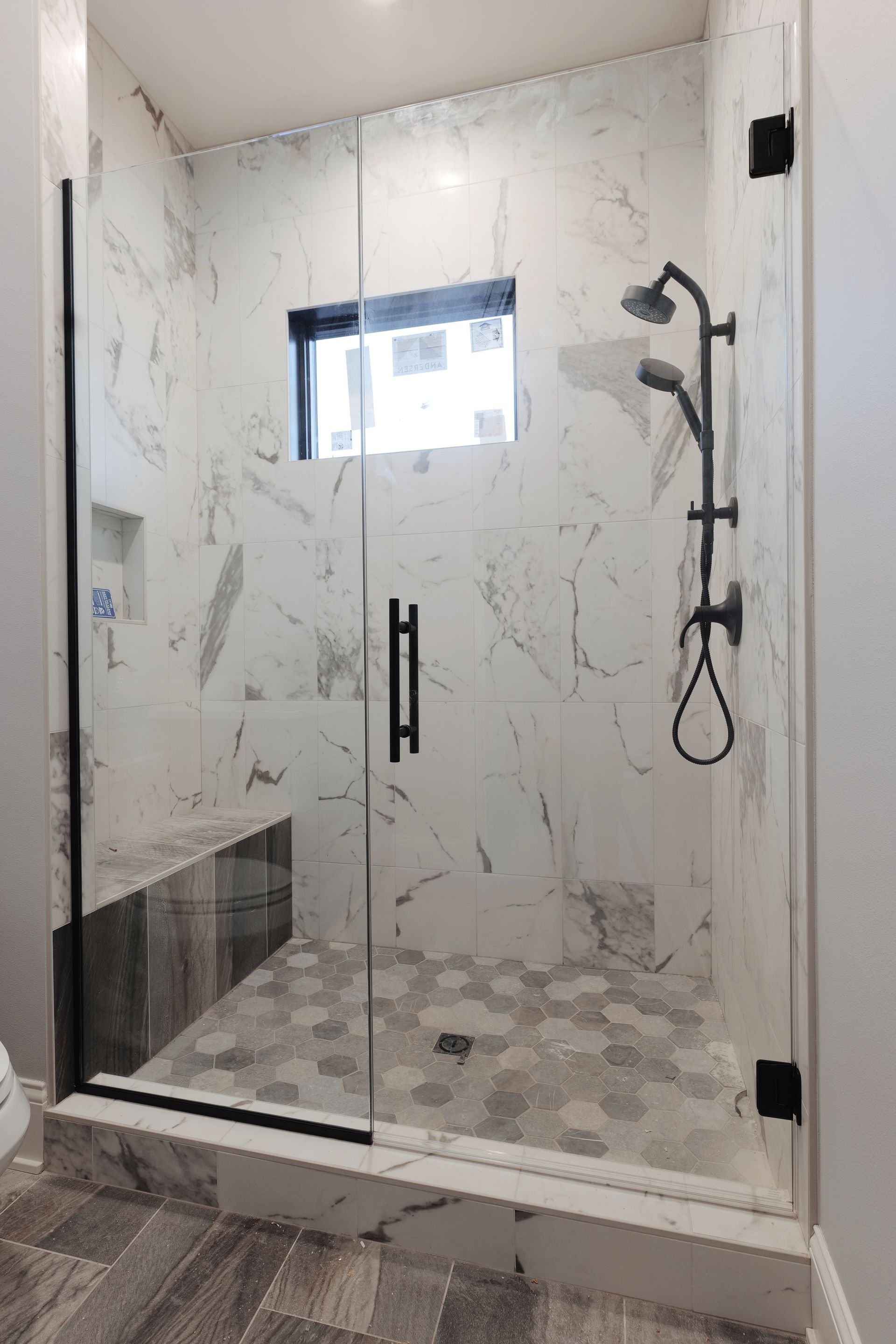 Modern bathroom with glass shower enclosure, marble walls, and gray hexagonal floor tiles.