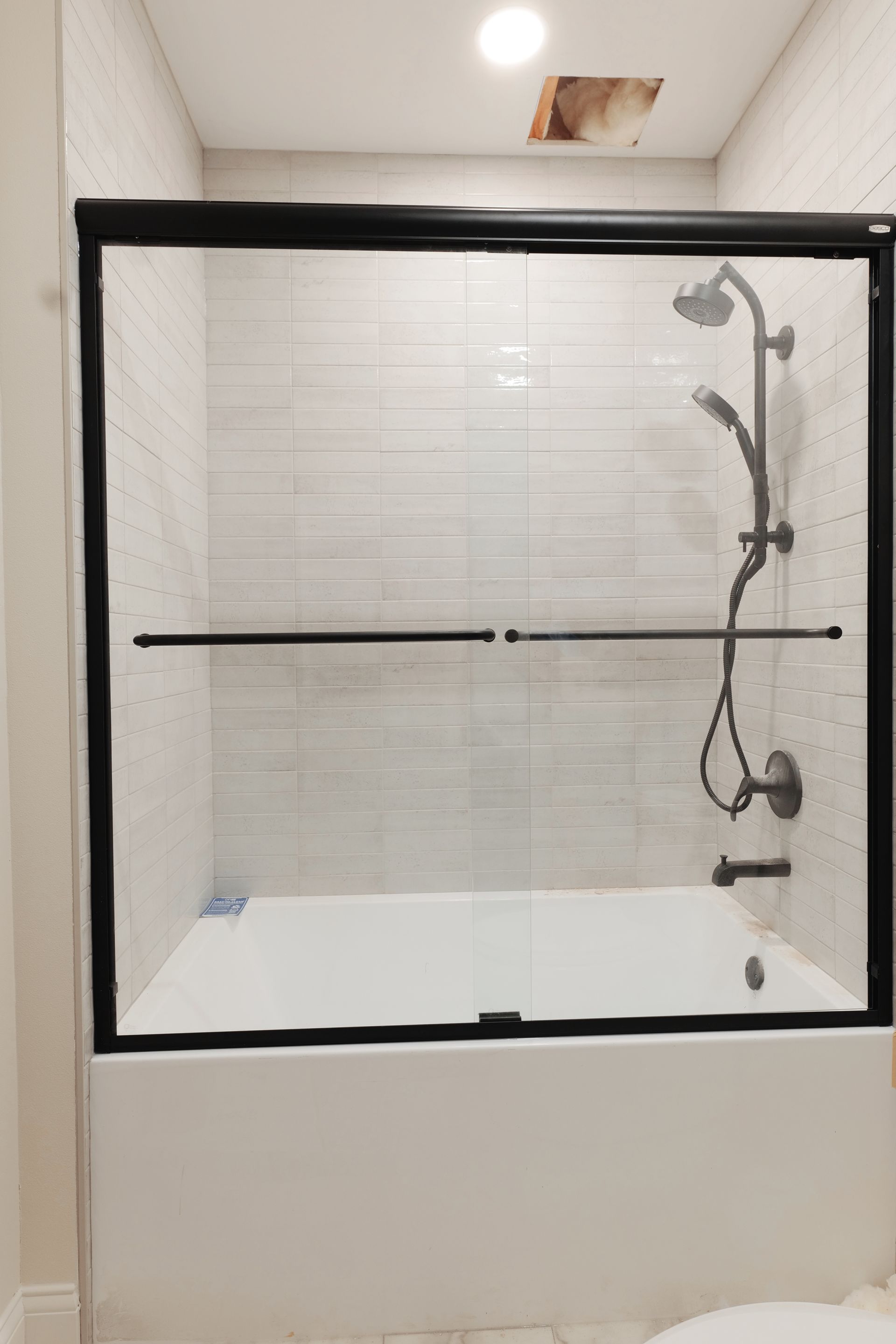 Black-framed glass shower door over a white tub with a dark shower head, set in a white-tiled bathroom.