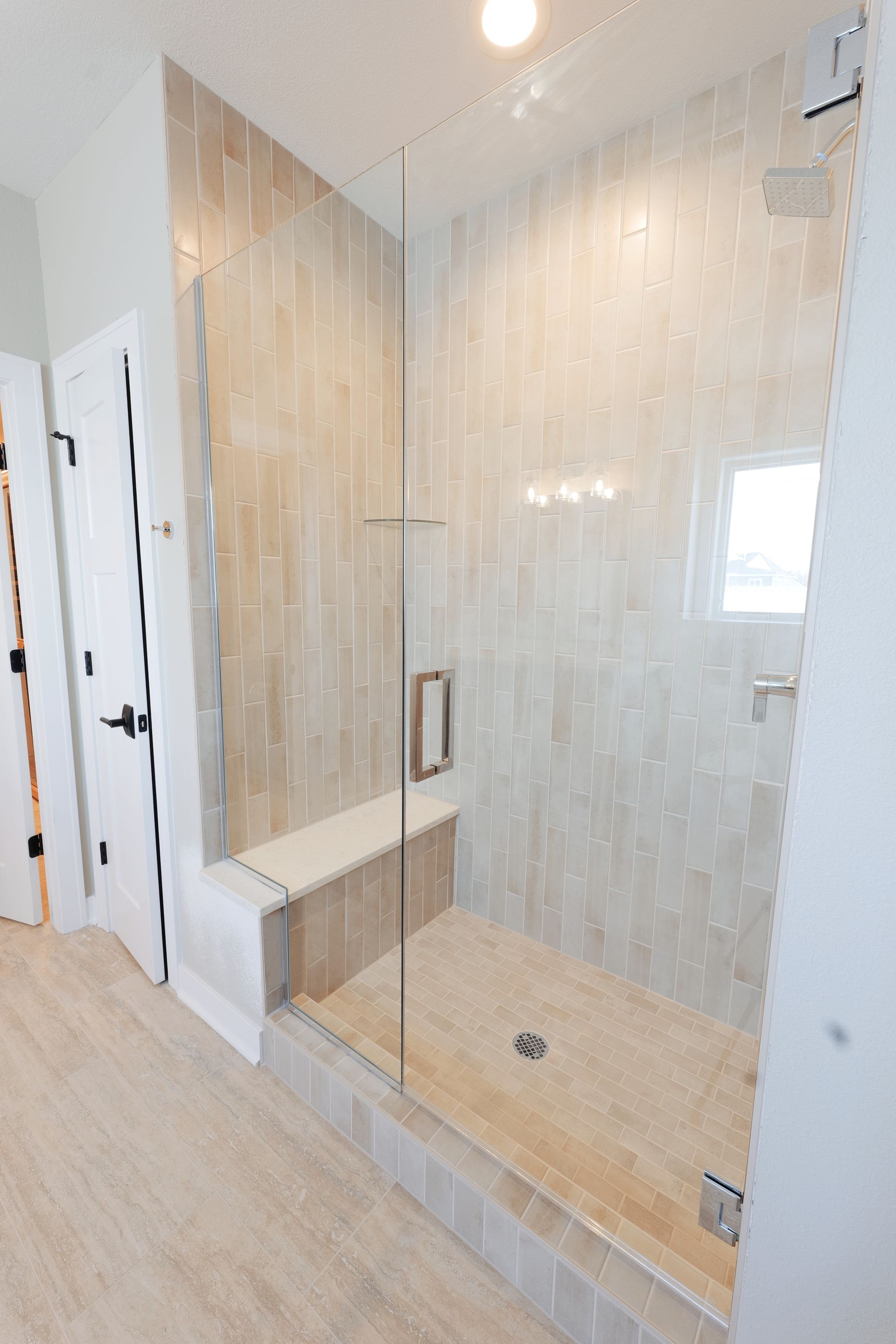 Glass-enclosed shower with tiled walls and a built-in bench in a light-colored bathroom.