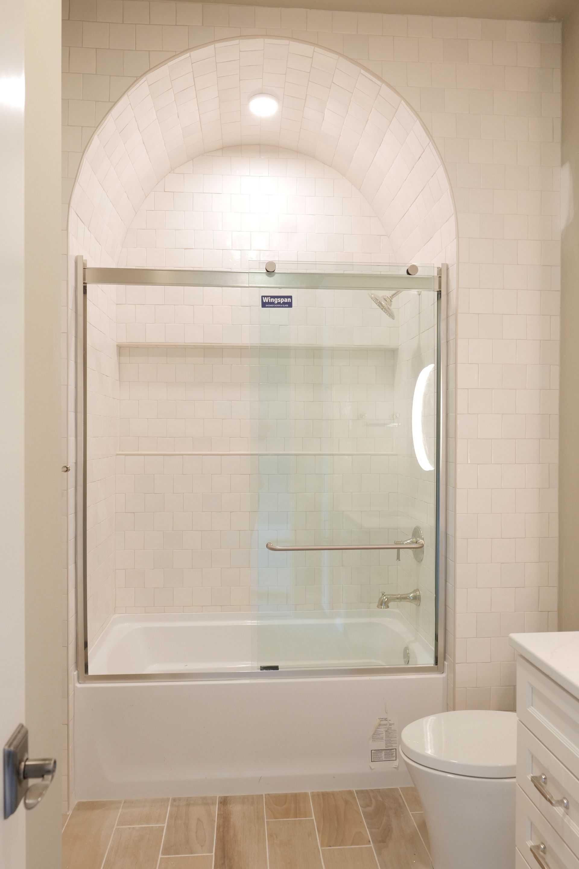 Bathroom with arched tile detail above a shower with sliding glass doors, white tub and toilet.