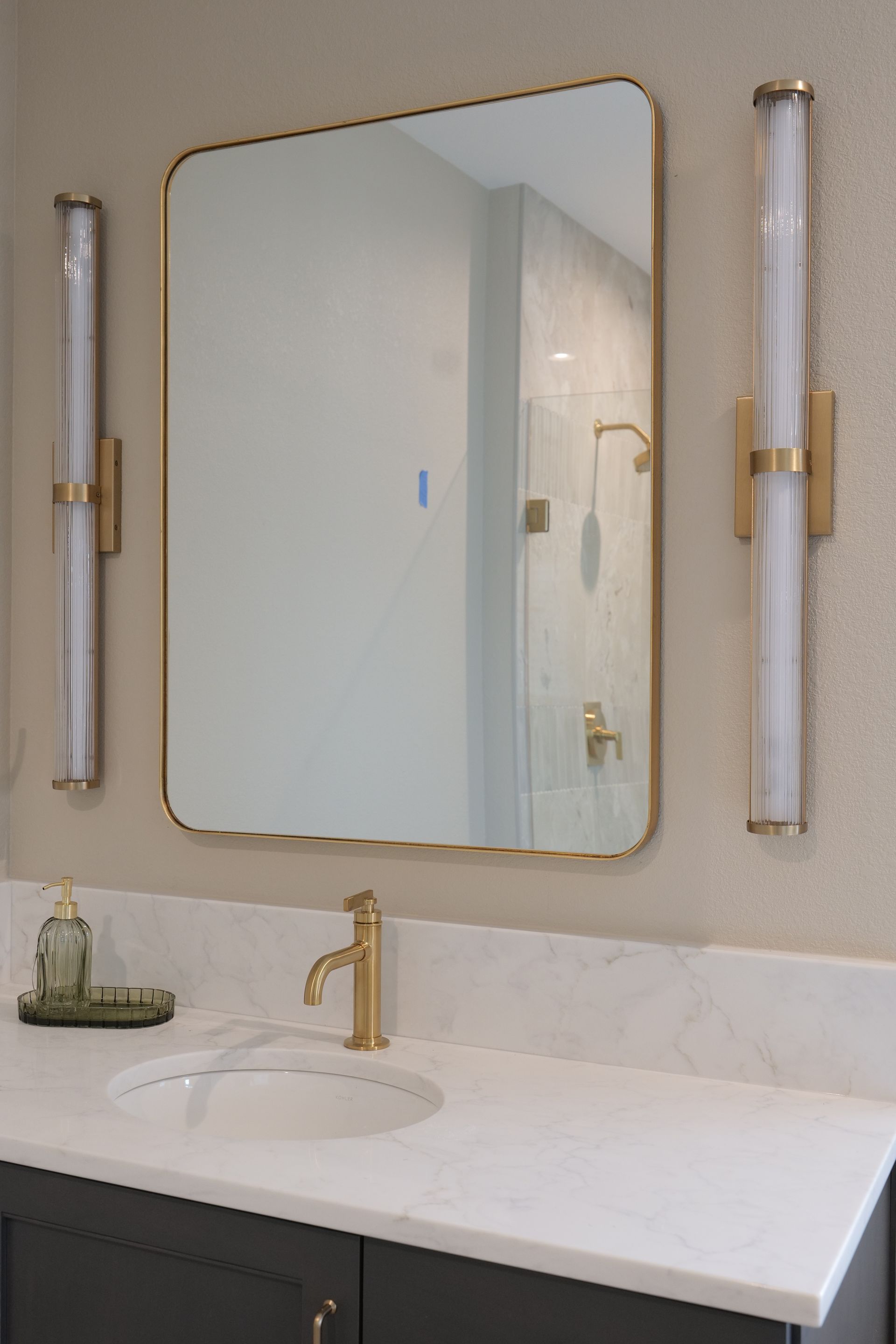Bathroom vanity with gold-framed mirror, marble countertop, gold faucet, and two vertical sconces.