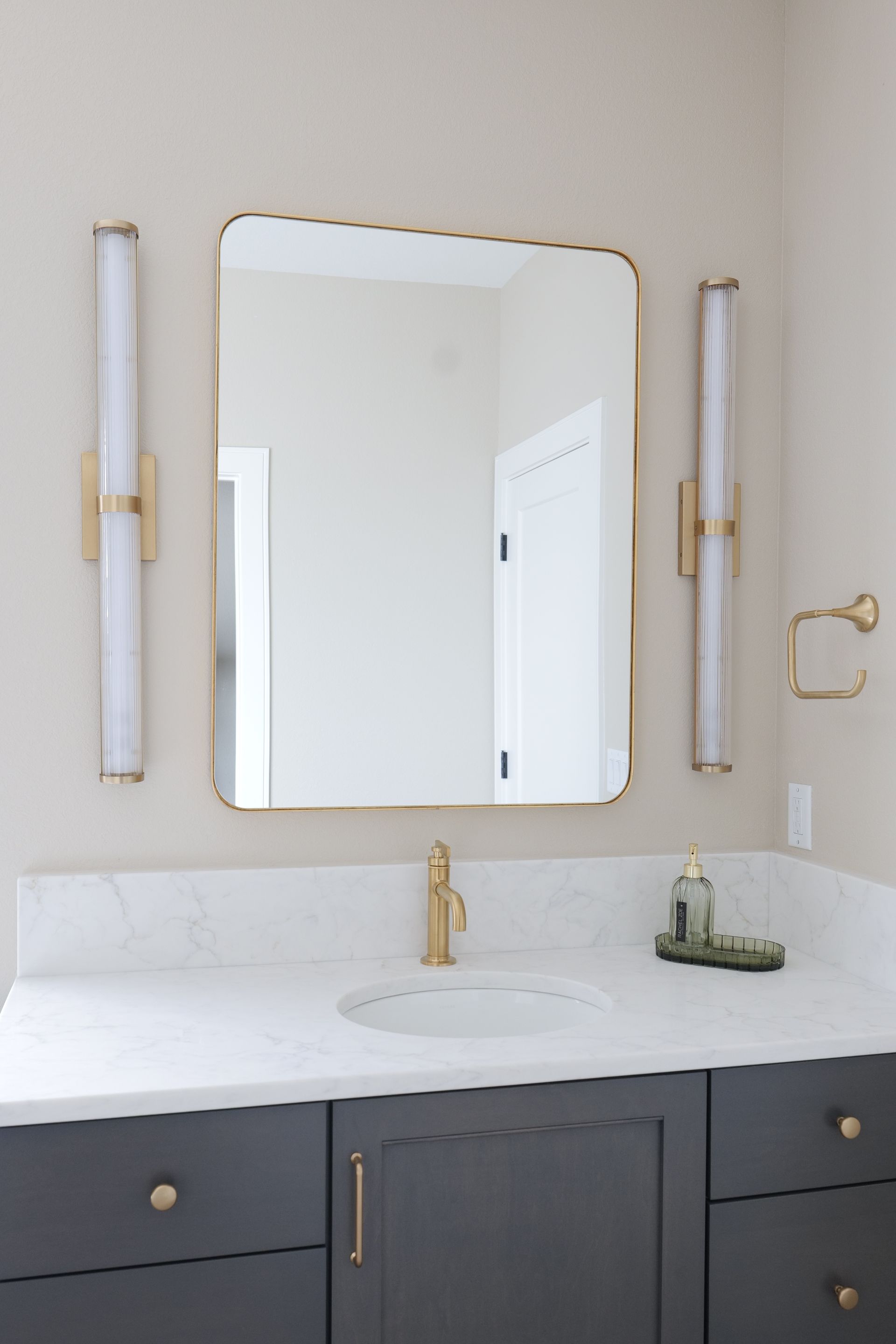 Bathroom vanity with gold-framed mirror, sconces, faucet, and dark gray cabinets with gold hardware.